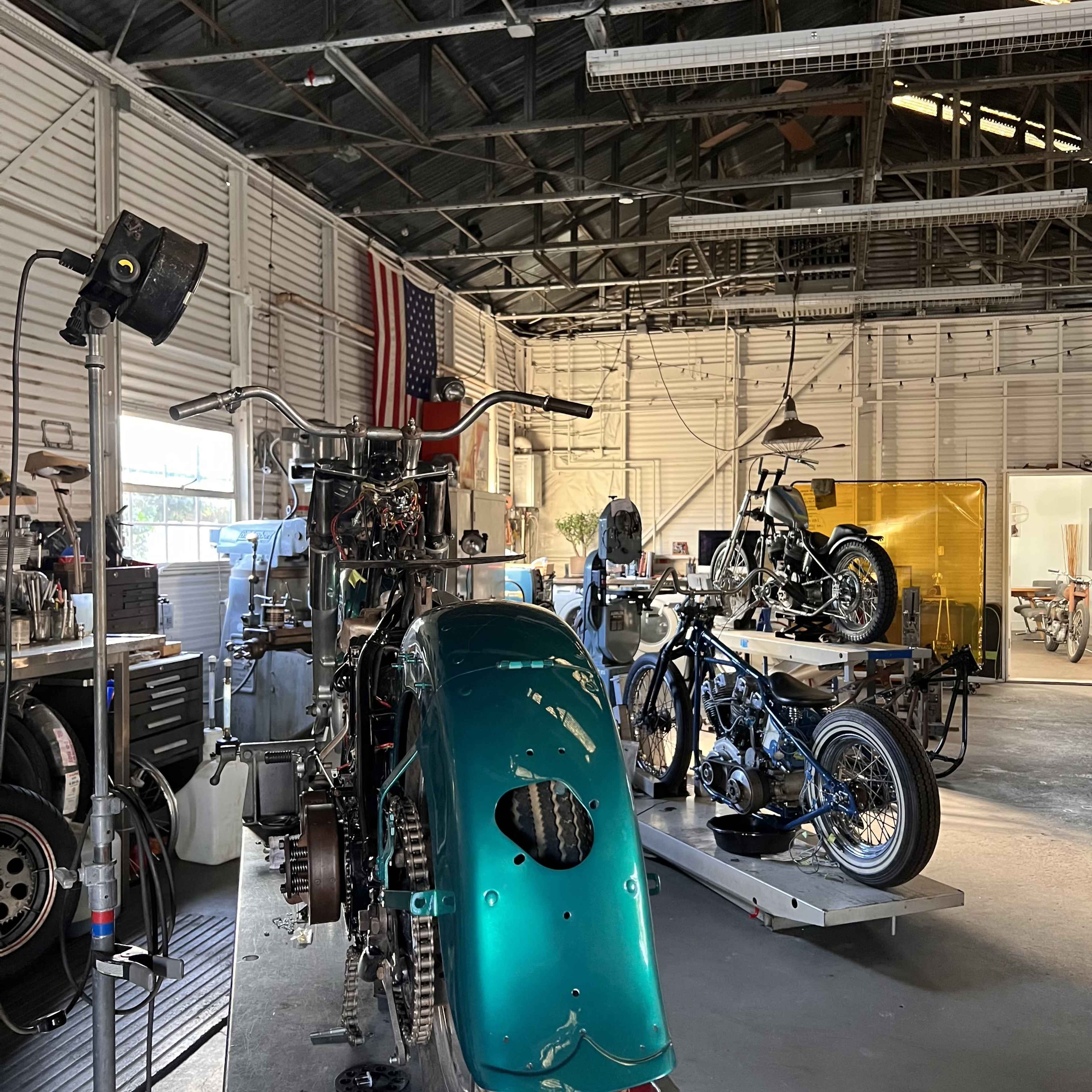 The image shows a motorcycle workshop with various motorcycle parts and two partially assembled motorcycles, set against a backdrop of bright lighting and an American flag.