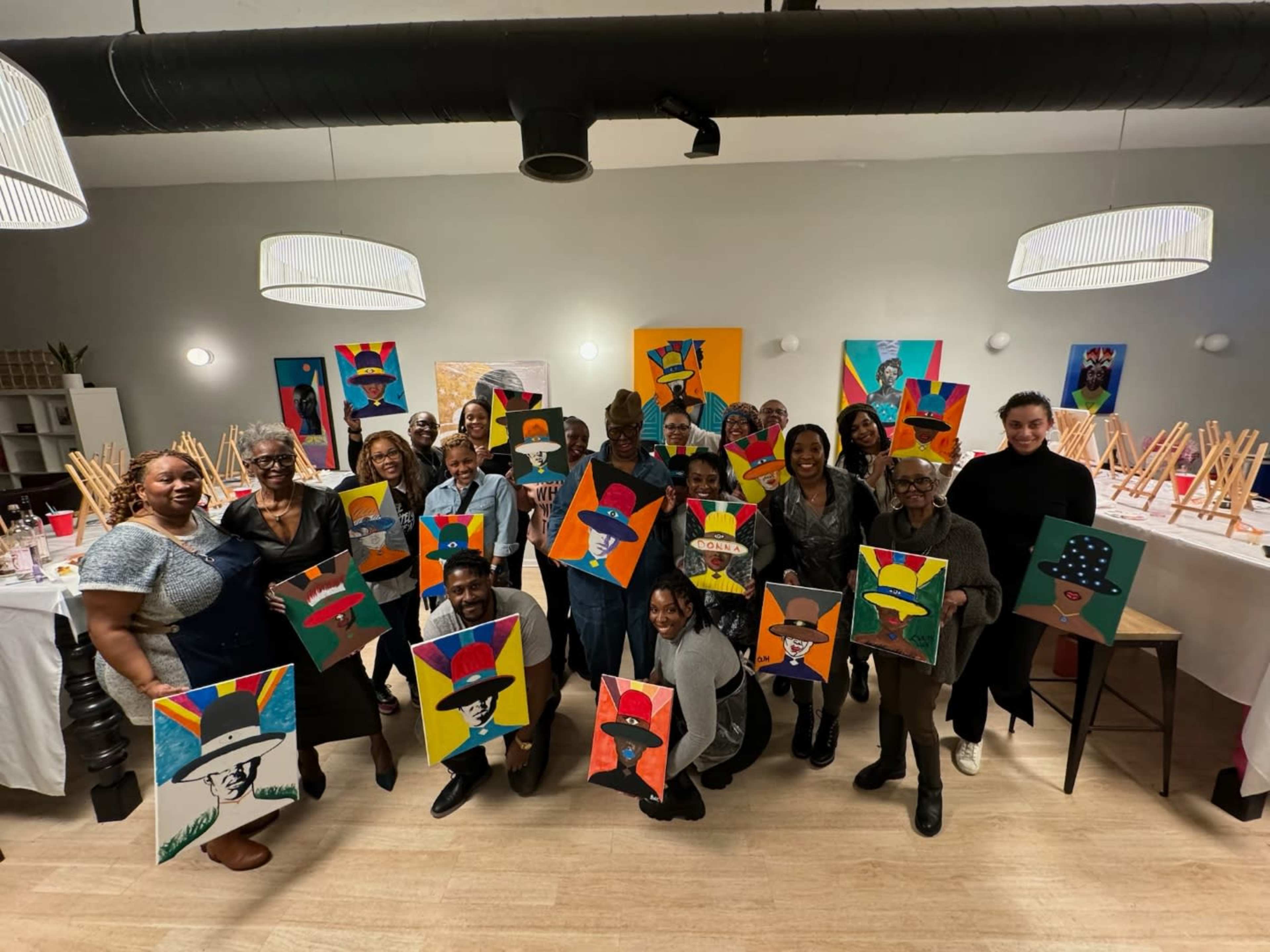 A diverse group of individuals holds paintings of colorful portraits in a studio setting adorned with easels and artwork in the background.