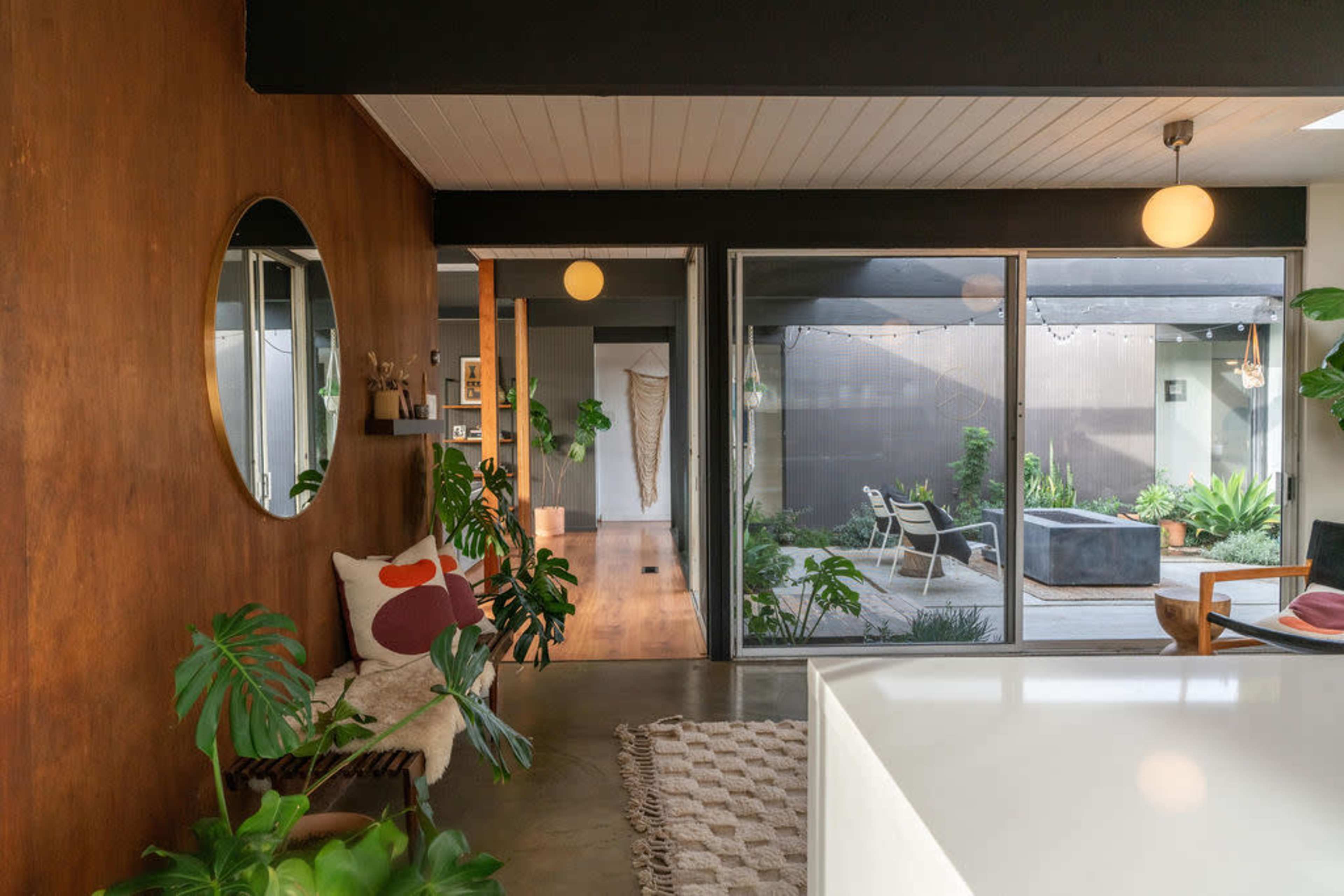 The image shows an interior space featuring a wooden wall, a round mirror, a sectional chair with a decorative pillow, and large glass doors leading to a landscaped outdoor area.