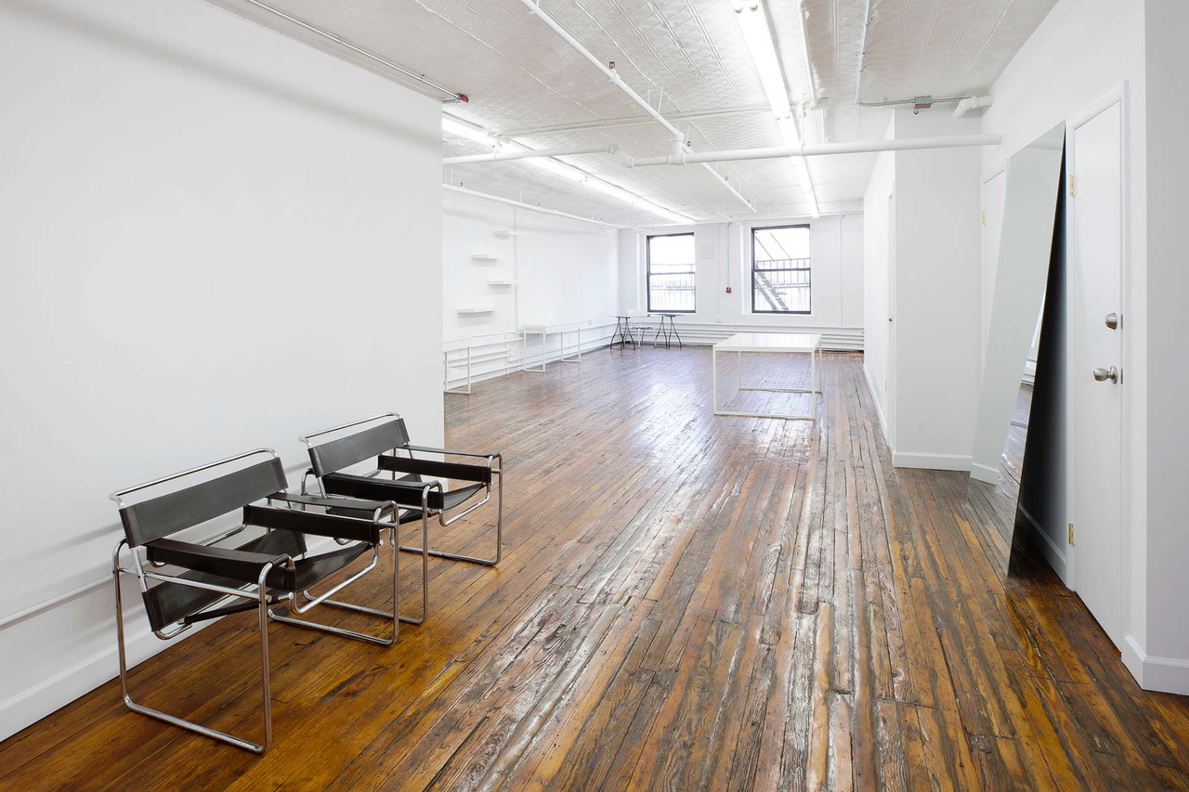 The image shows a spacious, empty room with wooden flooring, two black chairs near the wall, and minimalistic white walls.