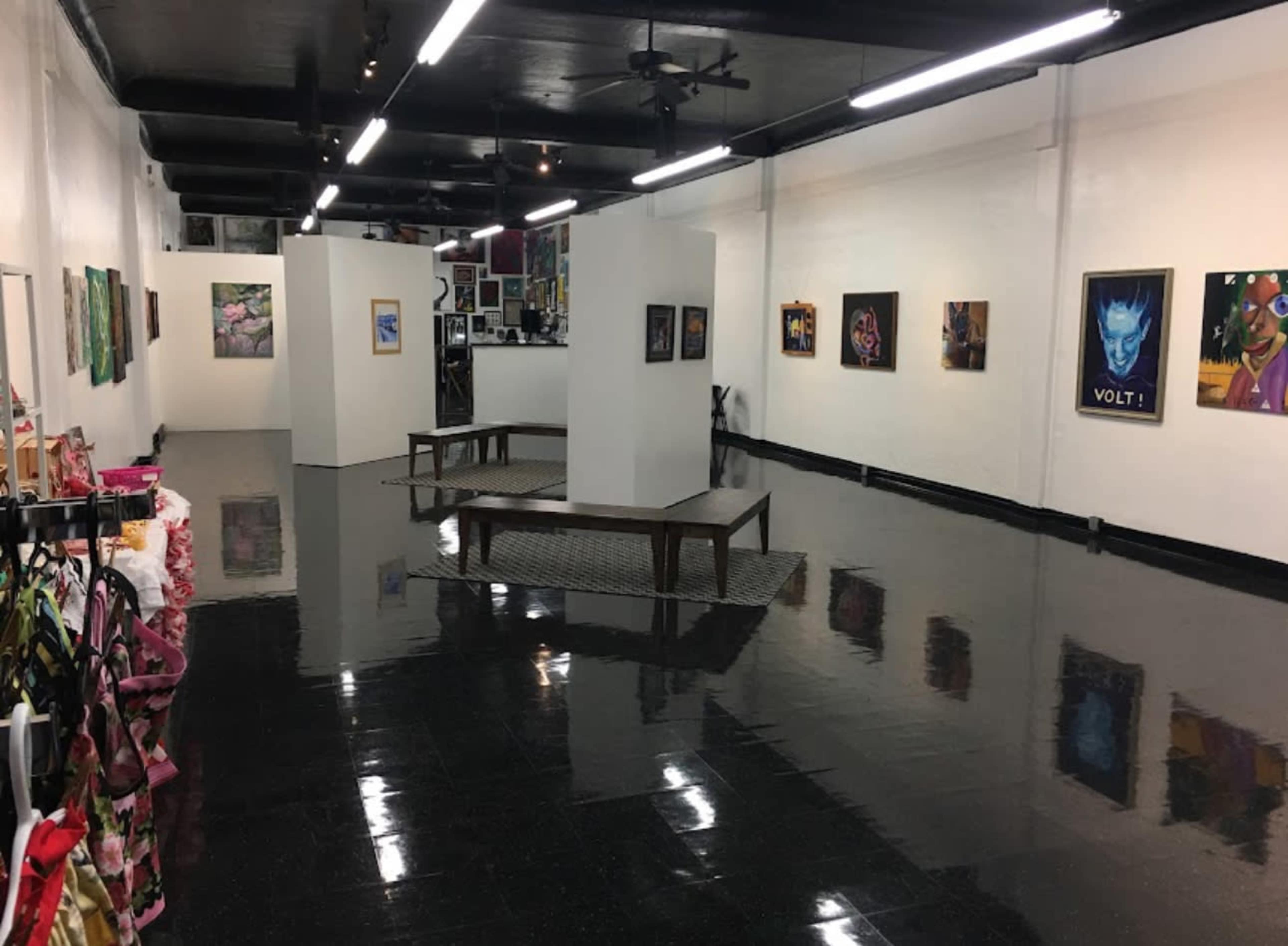 An art gallery displays various artworks on the walls, with white partitions and a central wooden platform in a glossy black-floored space.