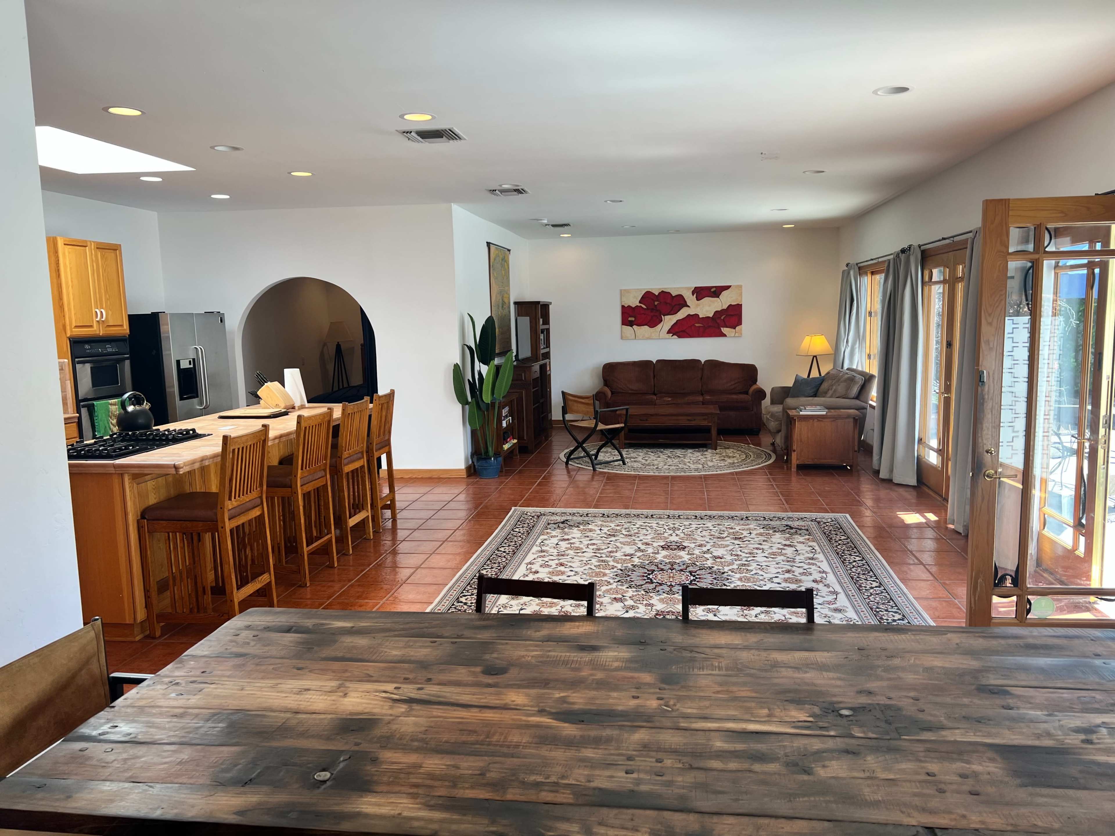 The image shows a spacious living area with a kitchen bar, dining table, and a cozy seating arrangement, featuring large windows that let in natural light.