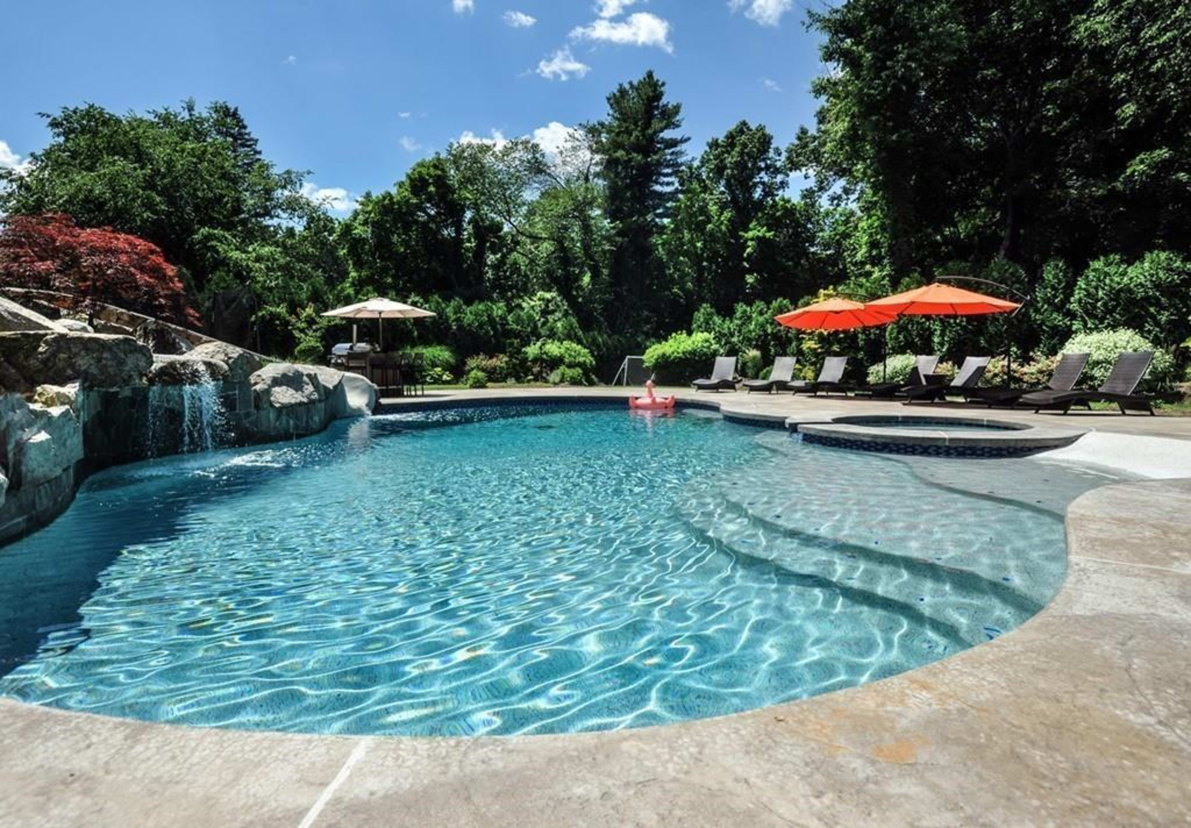 A swimming pool with a waterfall feature is surrounded by lounge chairs and umbrellas in a landscaped backyard.