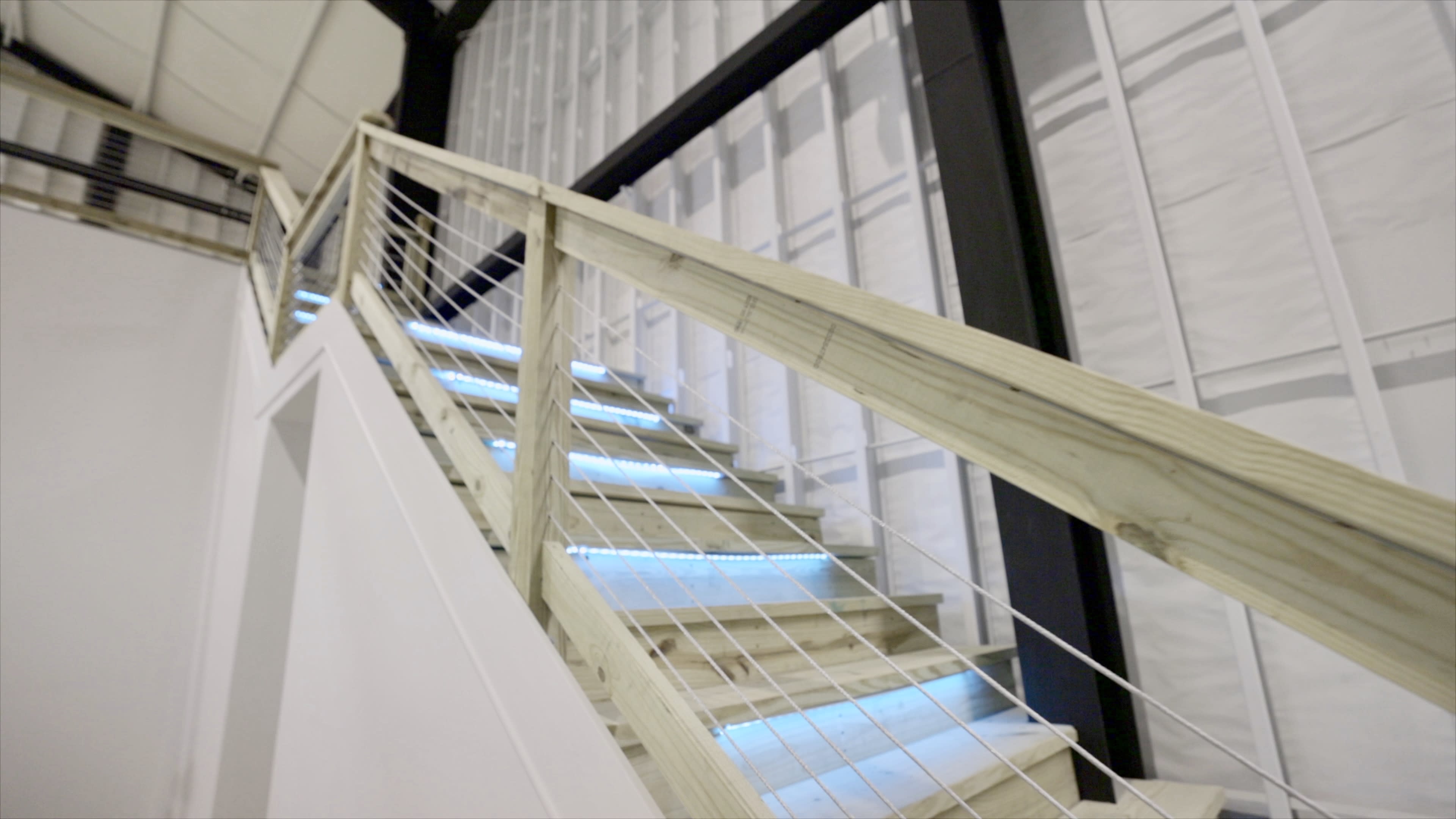 A wooden staircase with illuminated steps leading upward, surrounded by metal railings against a background of bare walls.