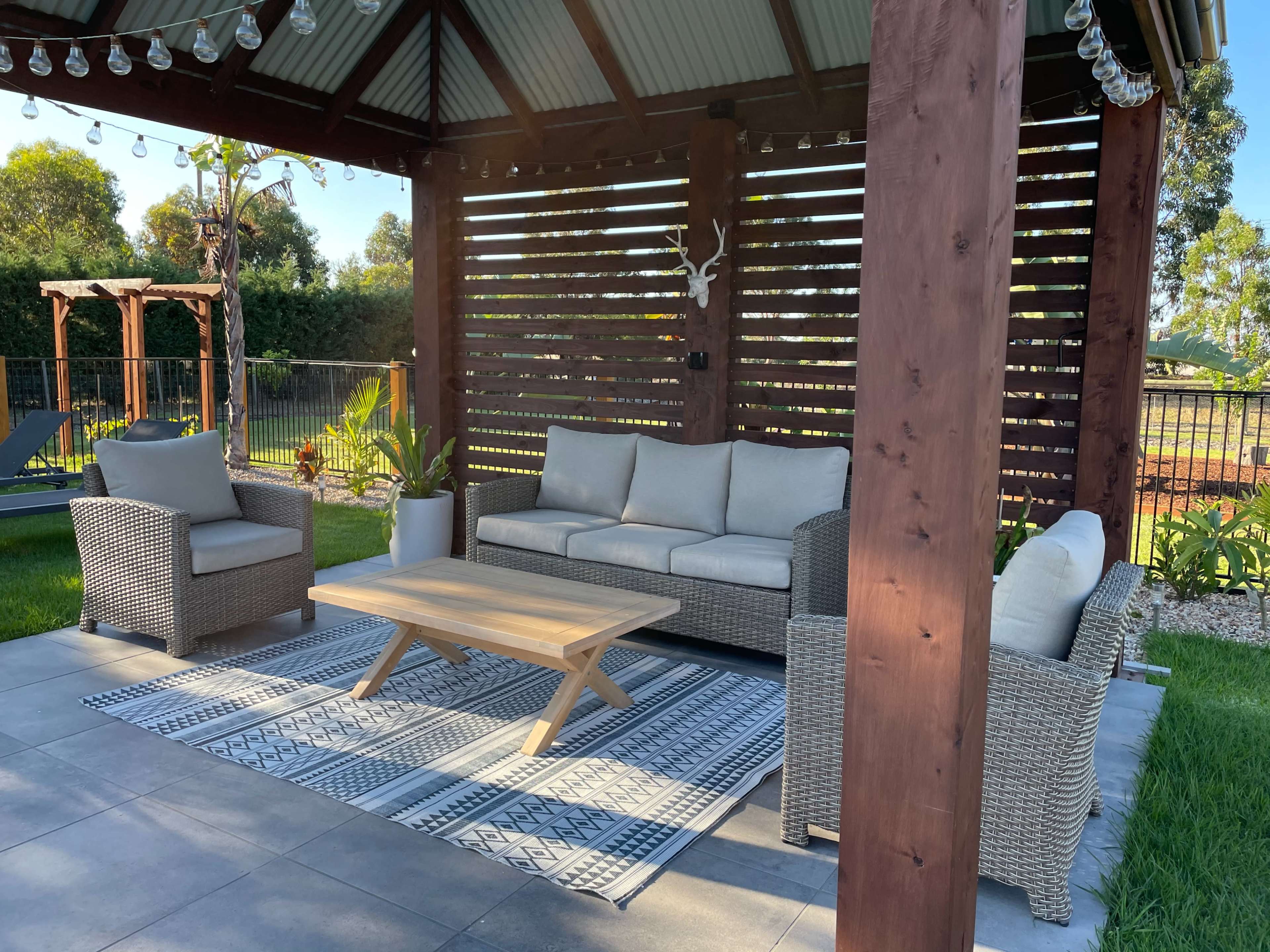 A covered outdoor seating area with a gray wicker sofa and two armchairs surrounding a wooden coffee table on a patterned rug.