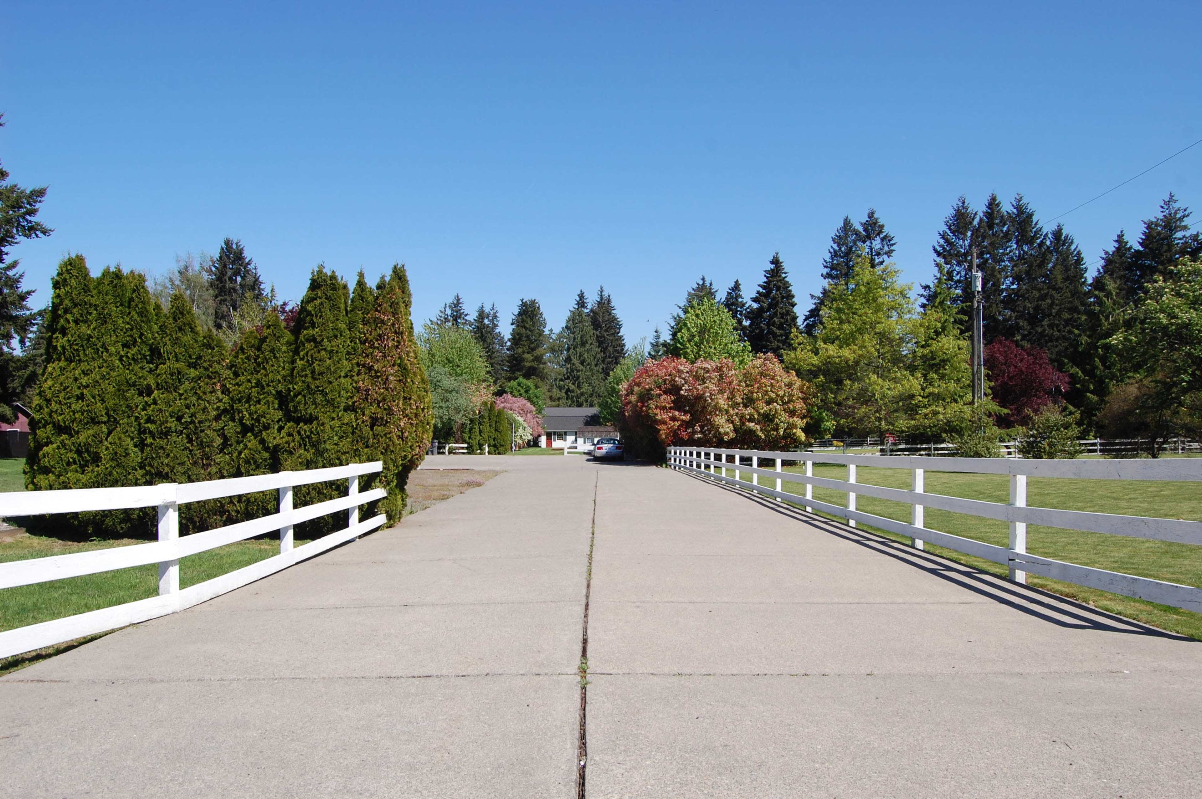 A paved path lined with white fences and greenery leads to a building at the end, surrounded by trees.