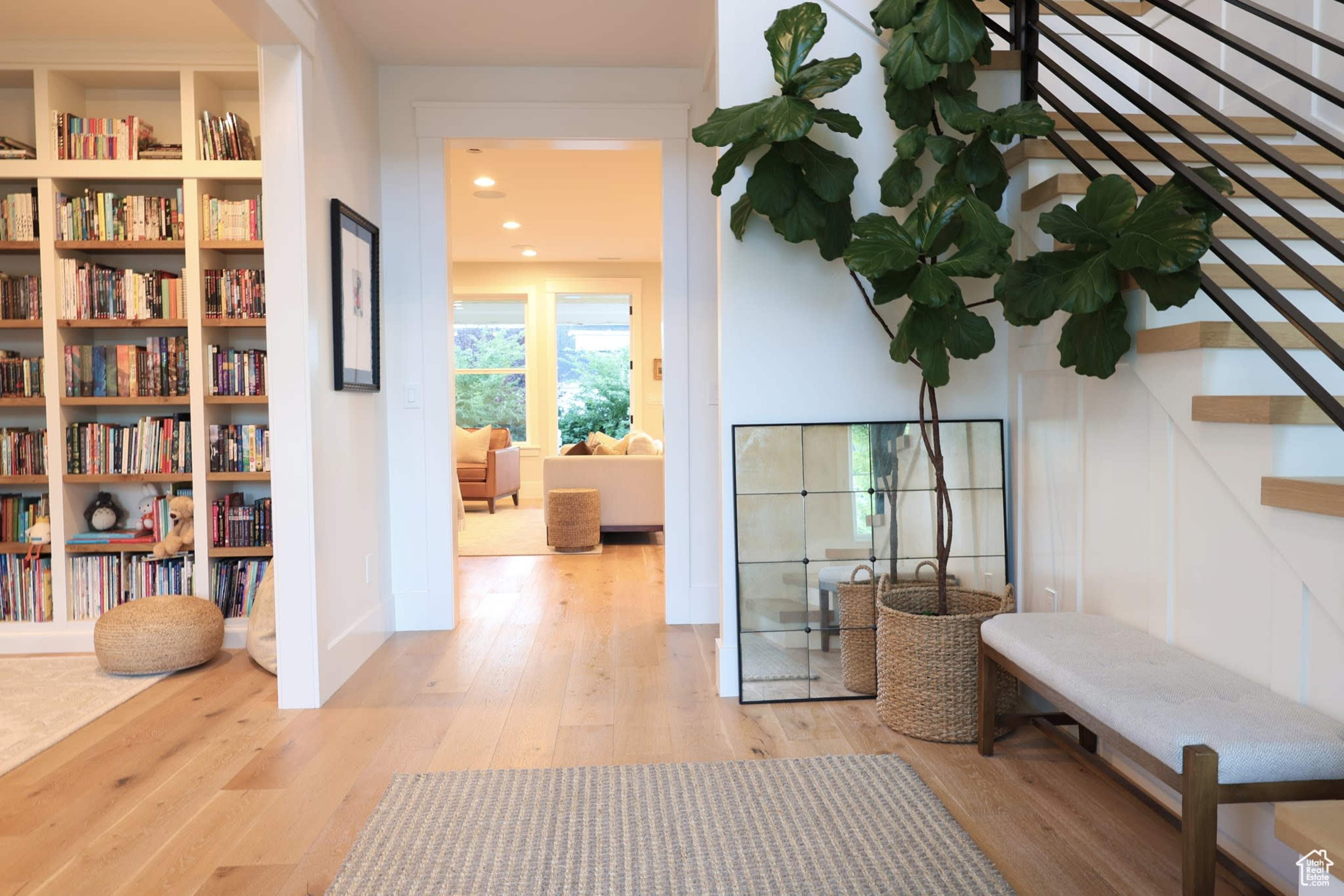 The image shows a bright and airy entryway featuring a bookshelf lined with books, a staircase with a metal railing, and a small seating area with a bench and decorative plants.