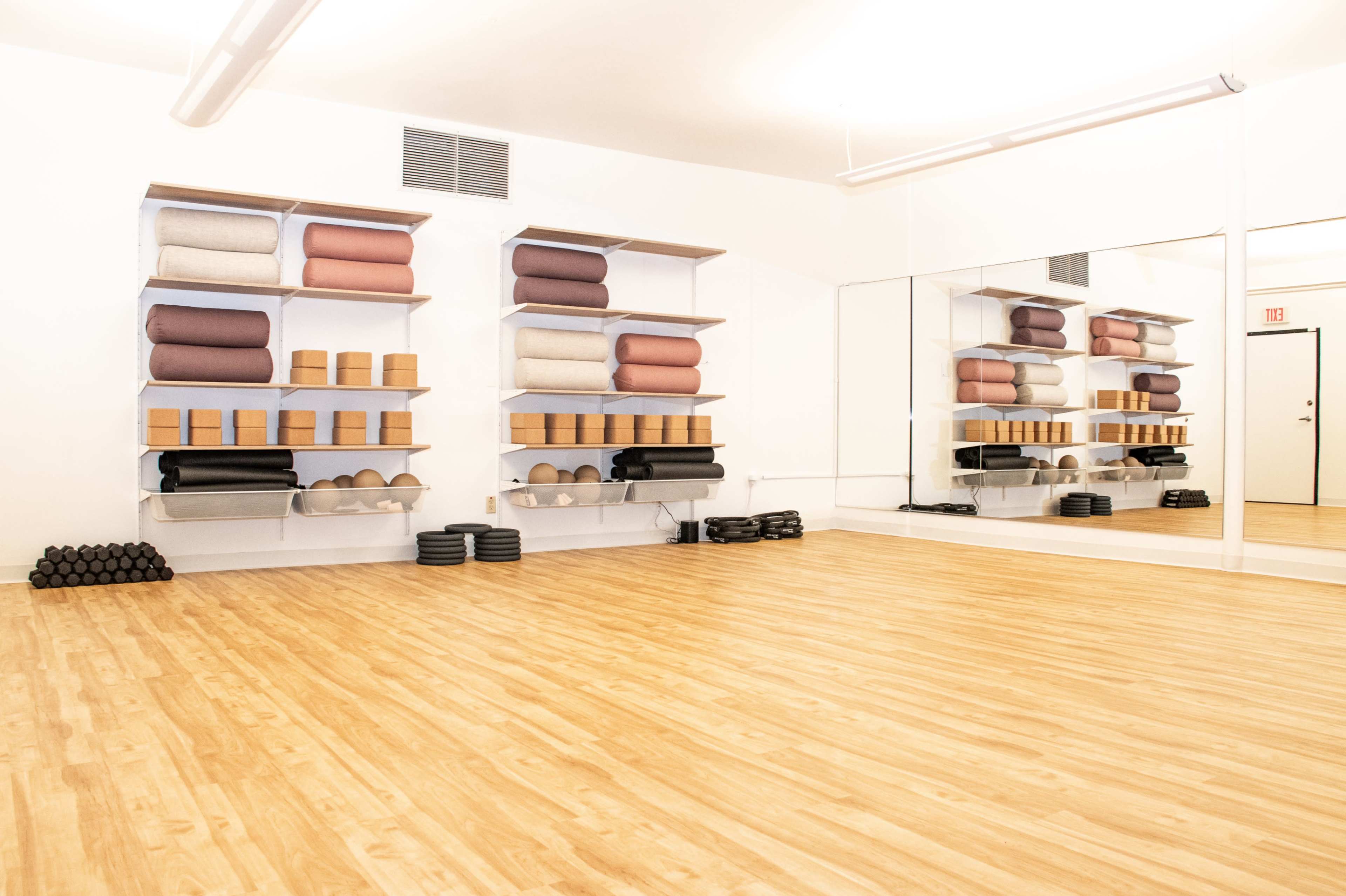 The image shows a yoga studio with wooden flooring, neatly organized shelves holding various yoga props, and a large mirror on one wall.