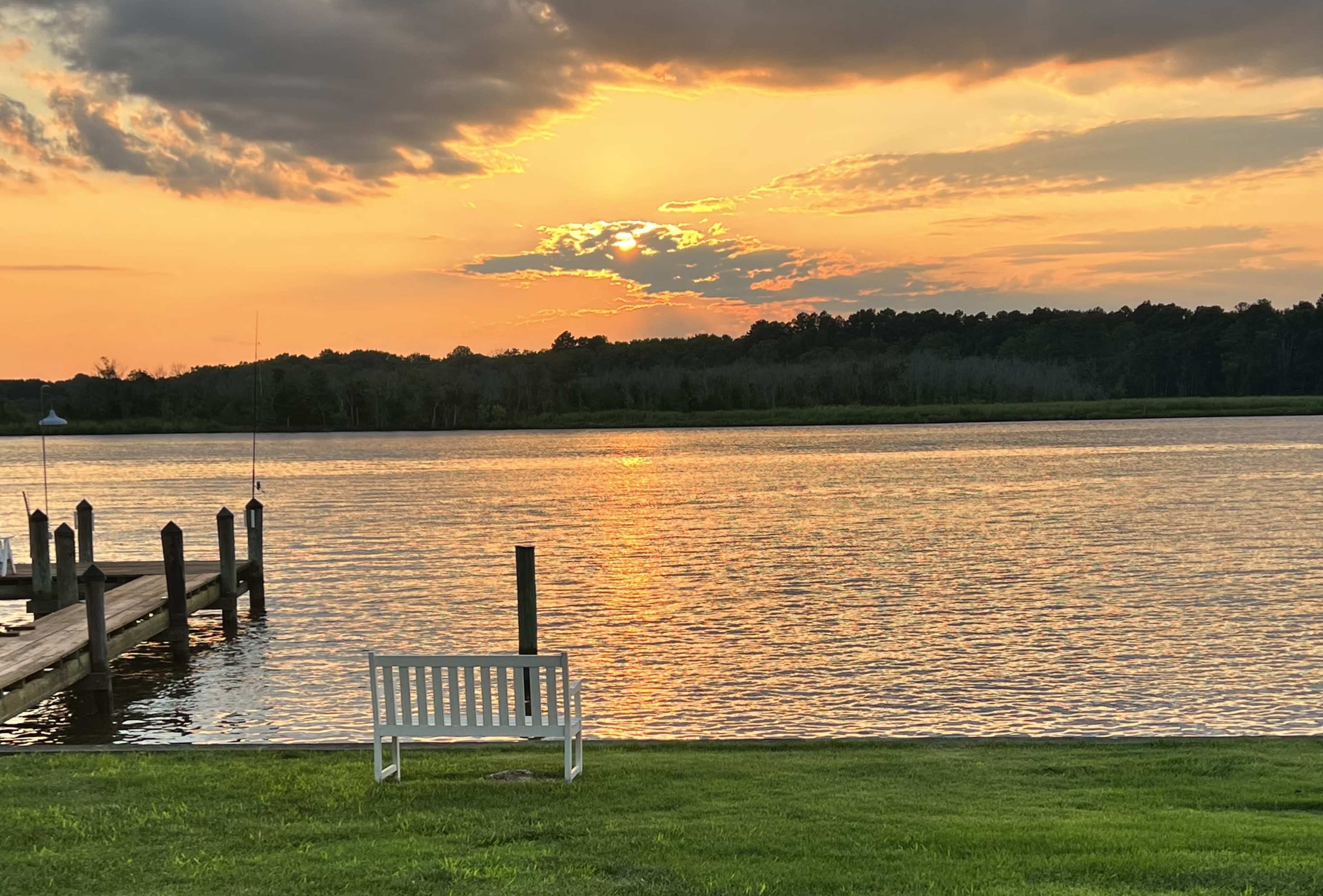 A white bench sits on a grassy area by a calm river, reflecting the colors of a sunset with clouds in the sky.