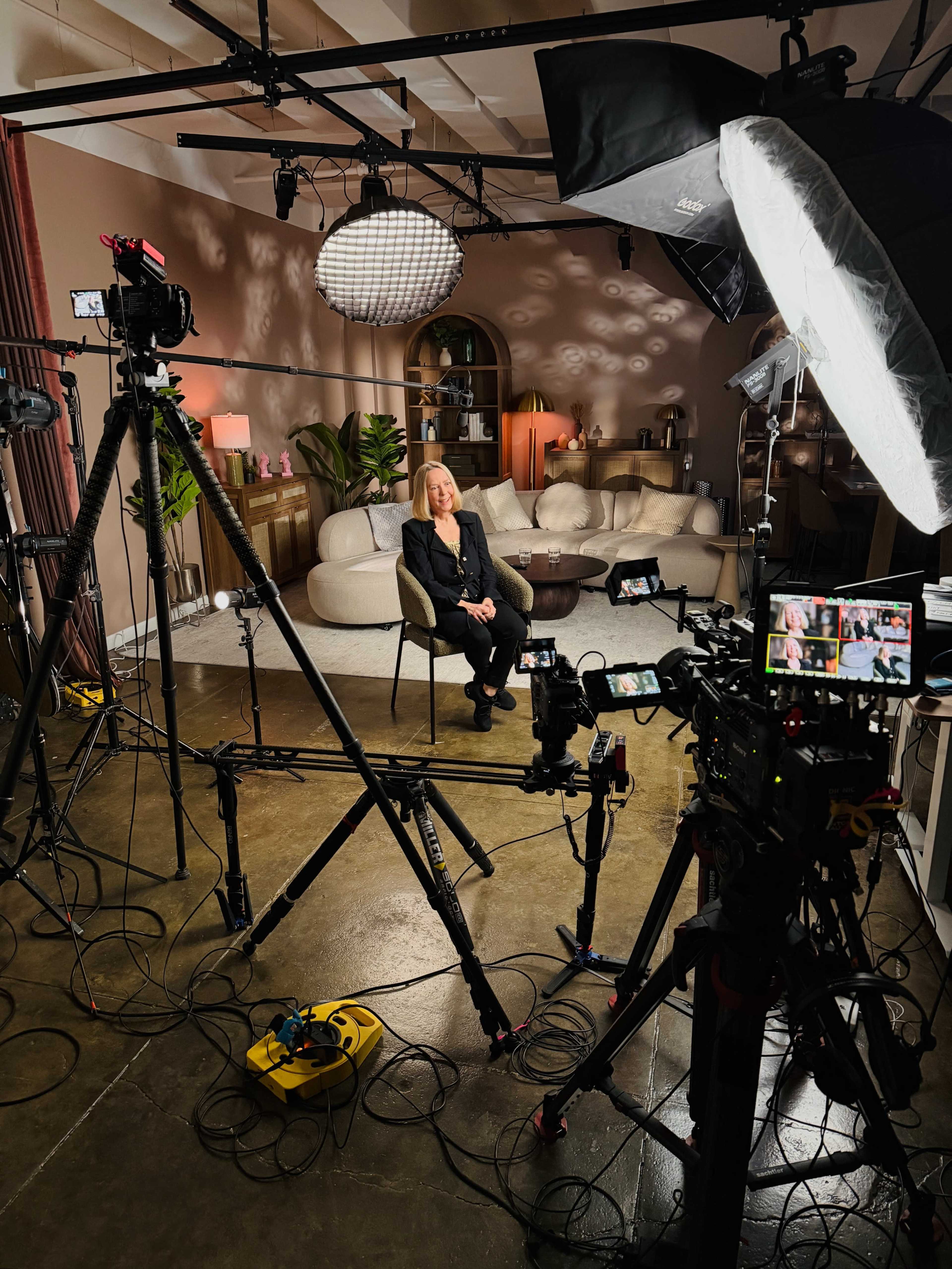 A woman sits on a chair in a well-lit studio surrounded by various cameras and lighting equipment.