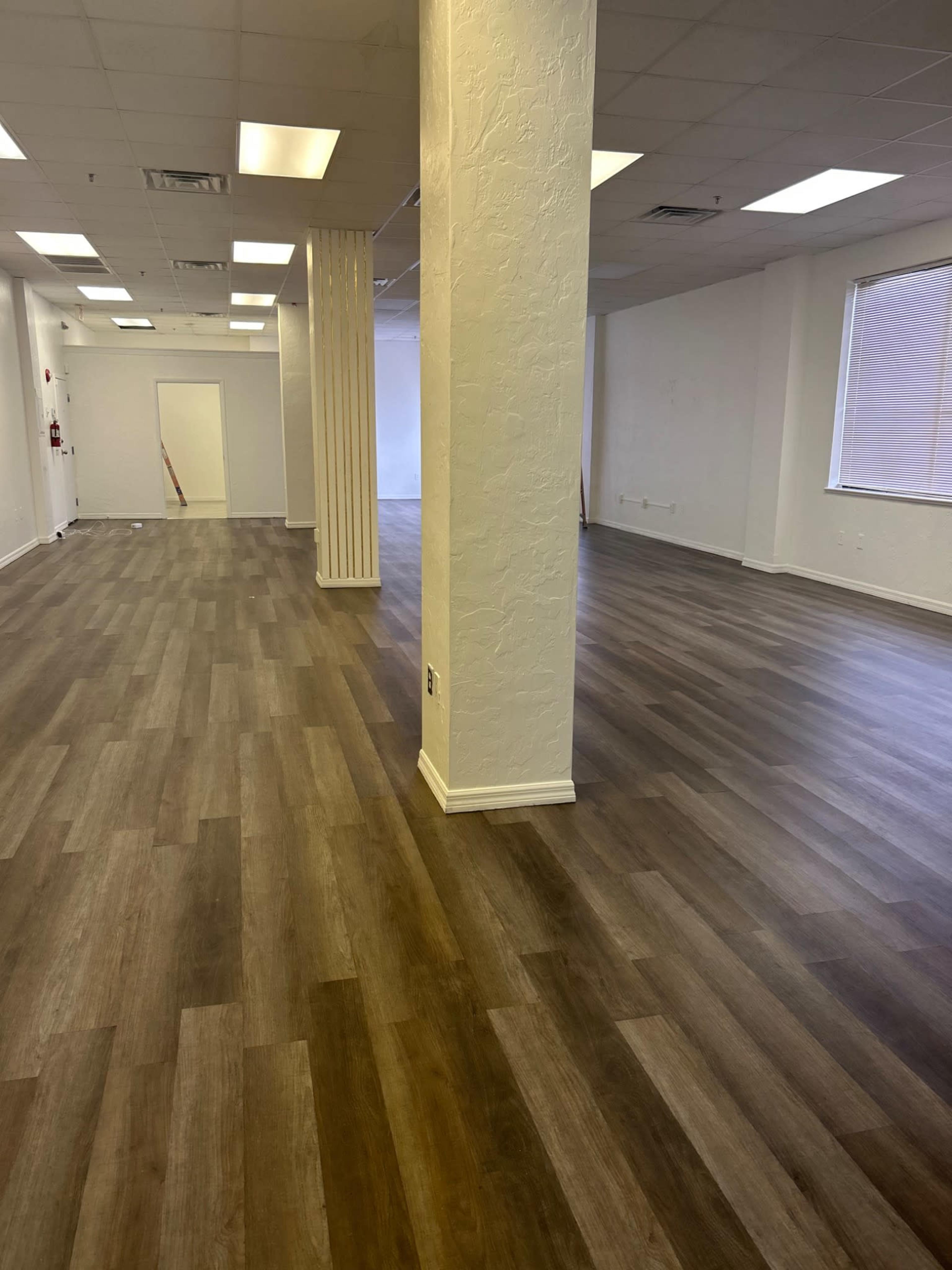 The image shows a spacious interior with light-colored walls, polished laminate flooring, and a column in the center.