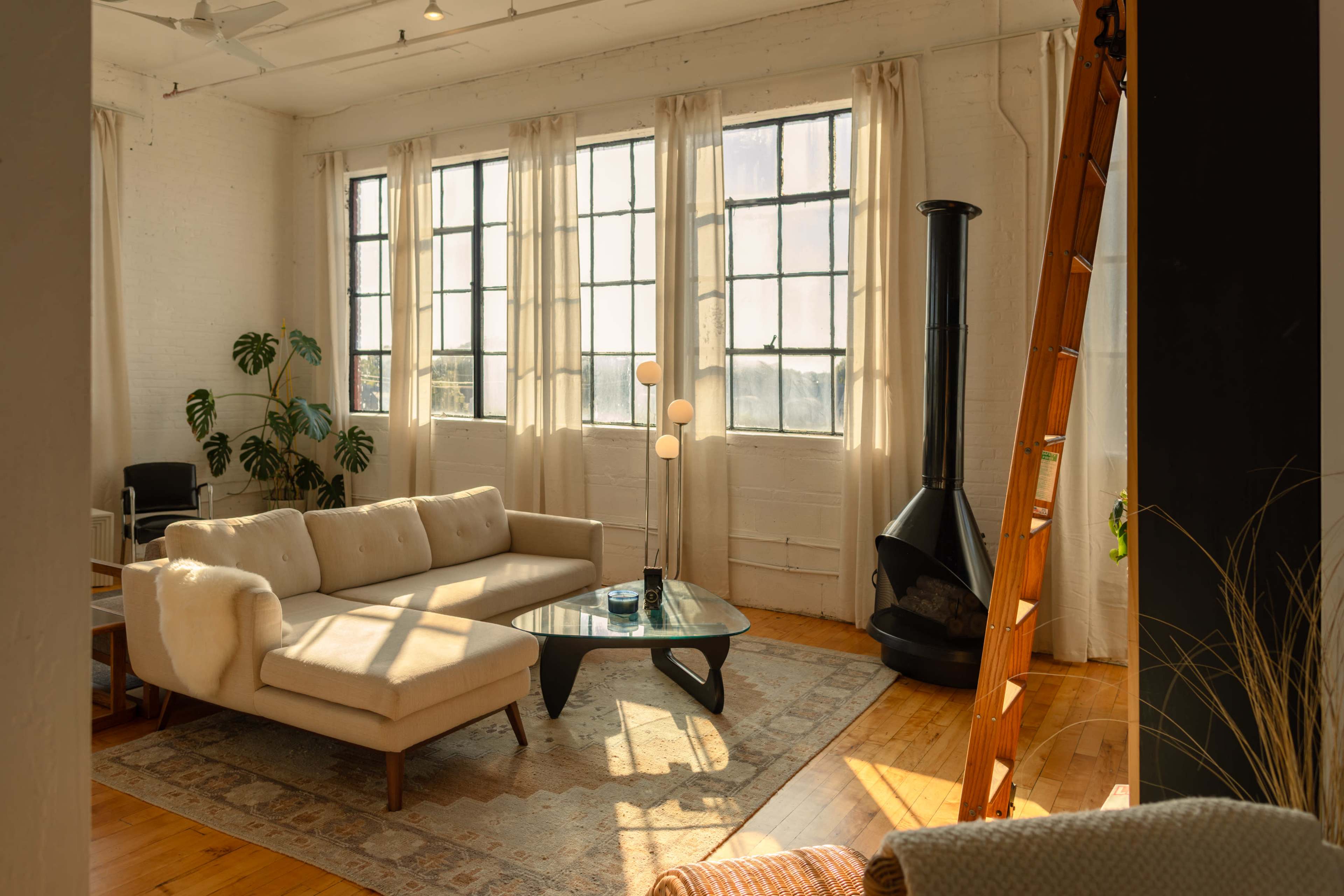The image shows a spacious, bright living room featuring a light-colored sofa, a glass coffee table, large windows with sheer curtains, and a modern wood-burning stove.