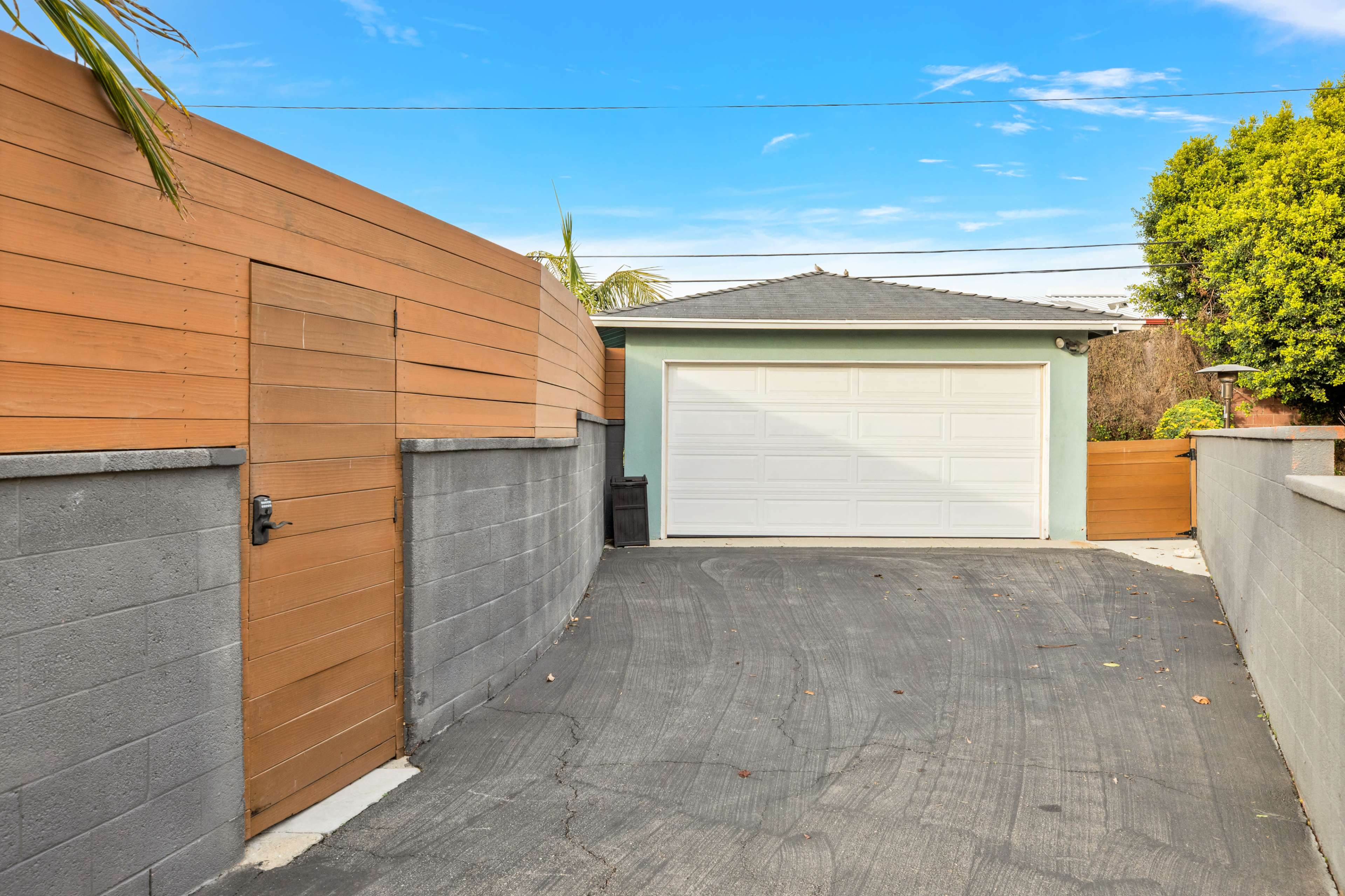 The image shows a driveway leading to a white garage door, bordered by wooden fencing and a concrete wall.