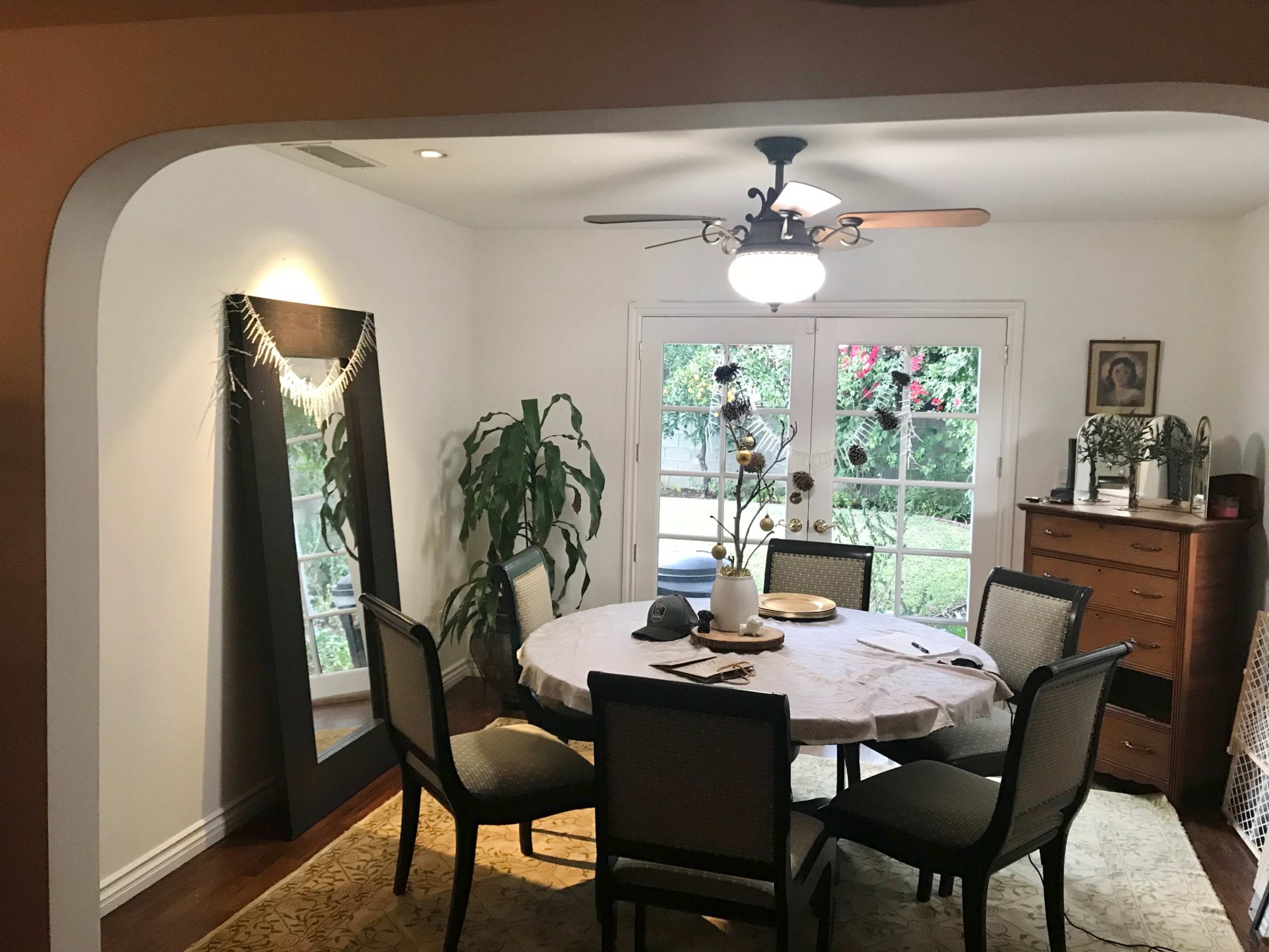 A dining room with a round table surrounded by six chairs, a mirror on the wall, and glass doors leading to a garden.