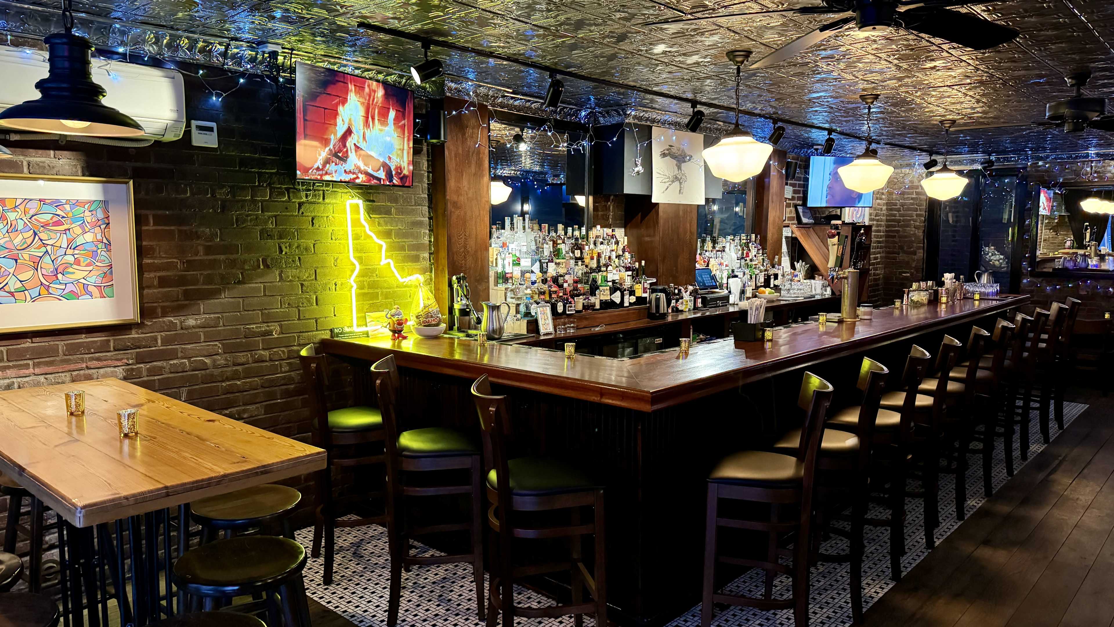 The image shows a dimly lit bar area featuring a long wooden bar lined with high stools, shelves stocked with various bottles, and illuminated by decorative lighting.