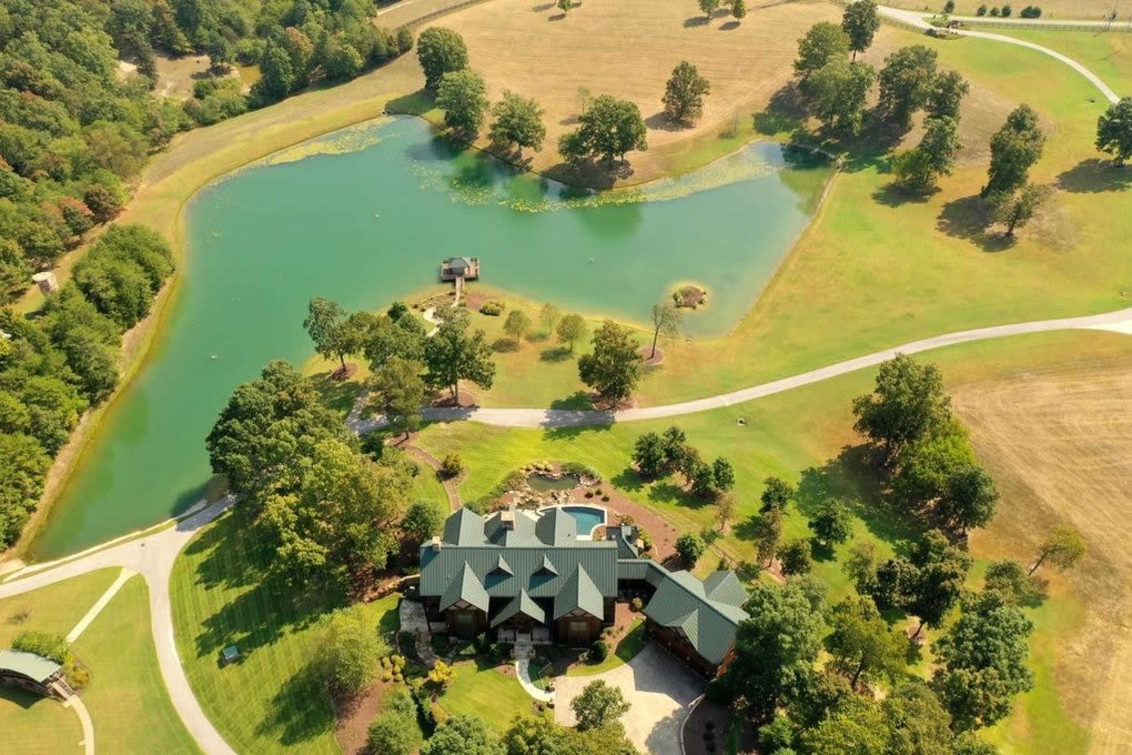 An aerial view shows a large, two-story house surrounded by green lawns, a pond, and several trees in a rural landscape.