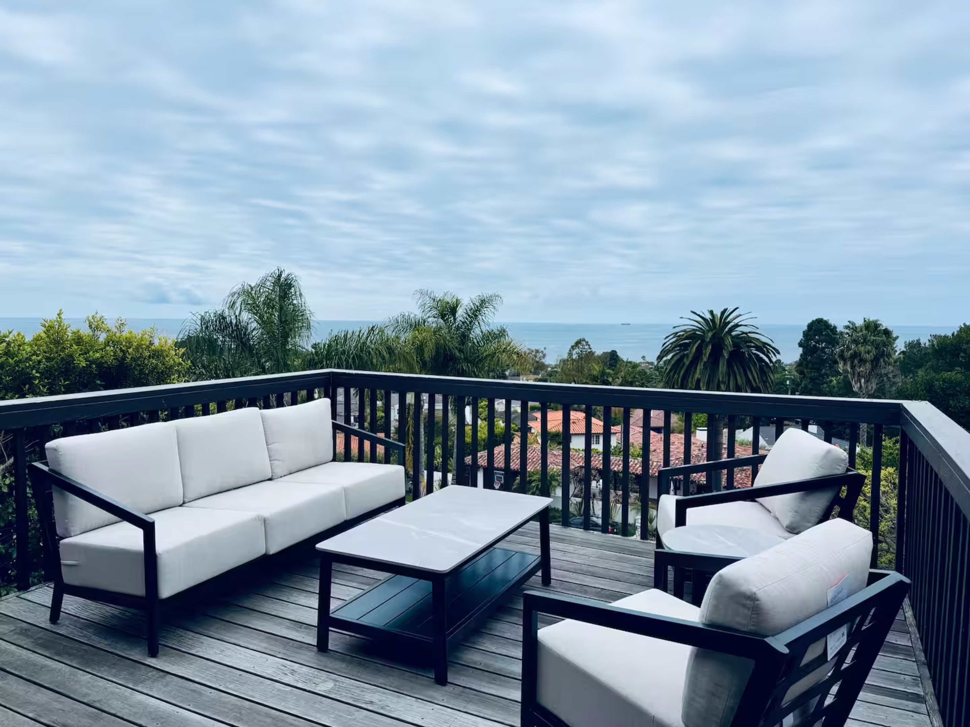 A wooden deck features a seating area with white sofas and a glass table, overlooking a view of the ocean and surrounding greenery.