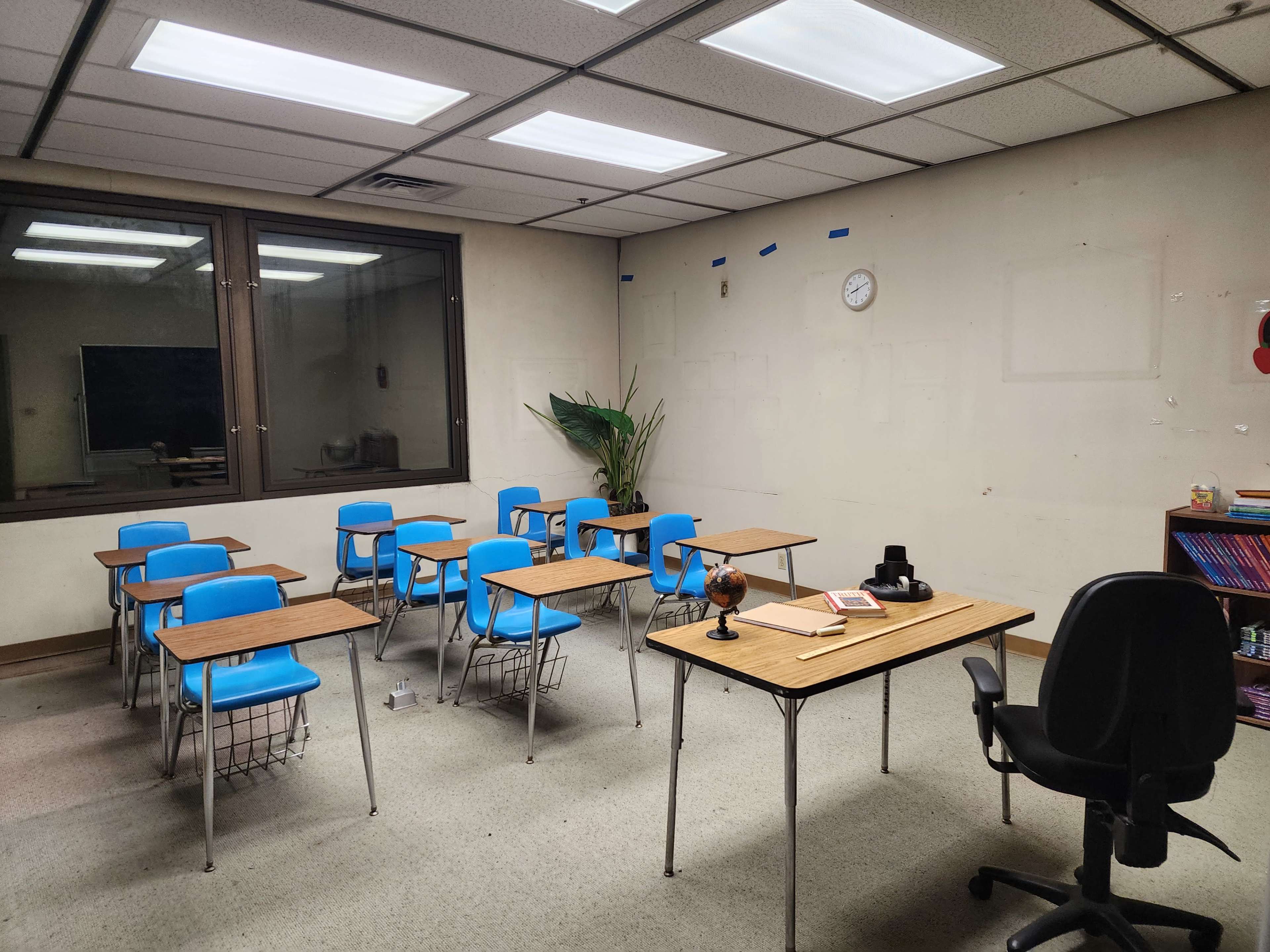 A sparsely furnished classroom with blue chairs, wooden tables, a globe, and a large window.