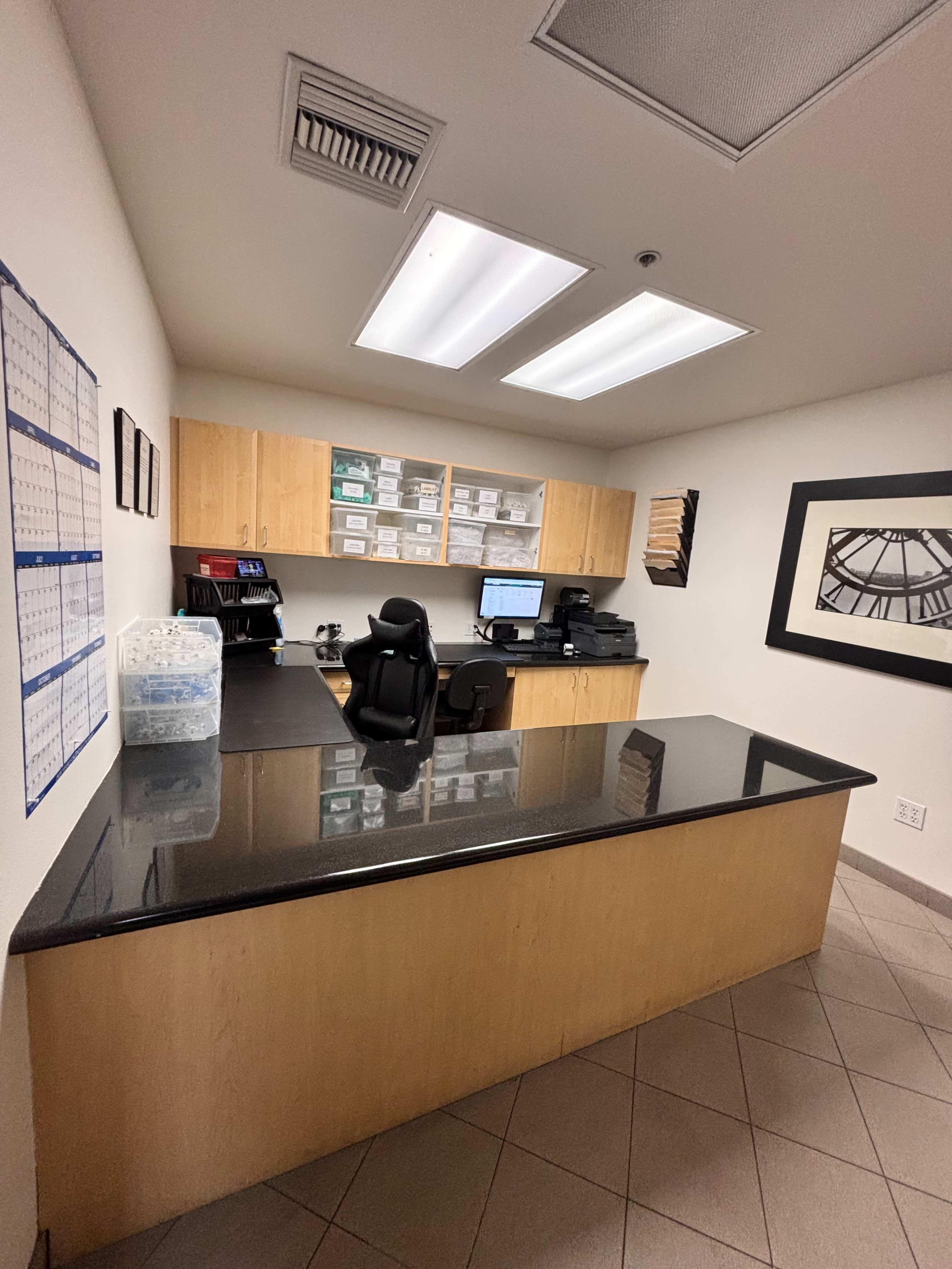 The image shows a neat office space featuring a reception desk with a computer, printer, and storage cabinets, alongside a calendar on the wall.