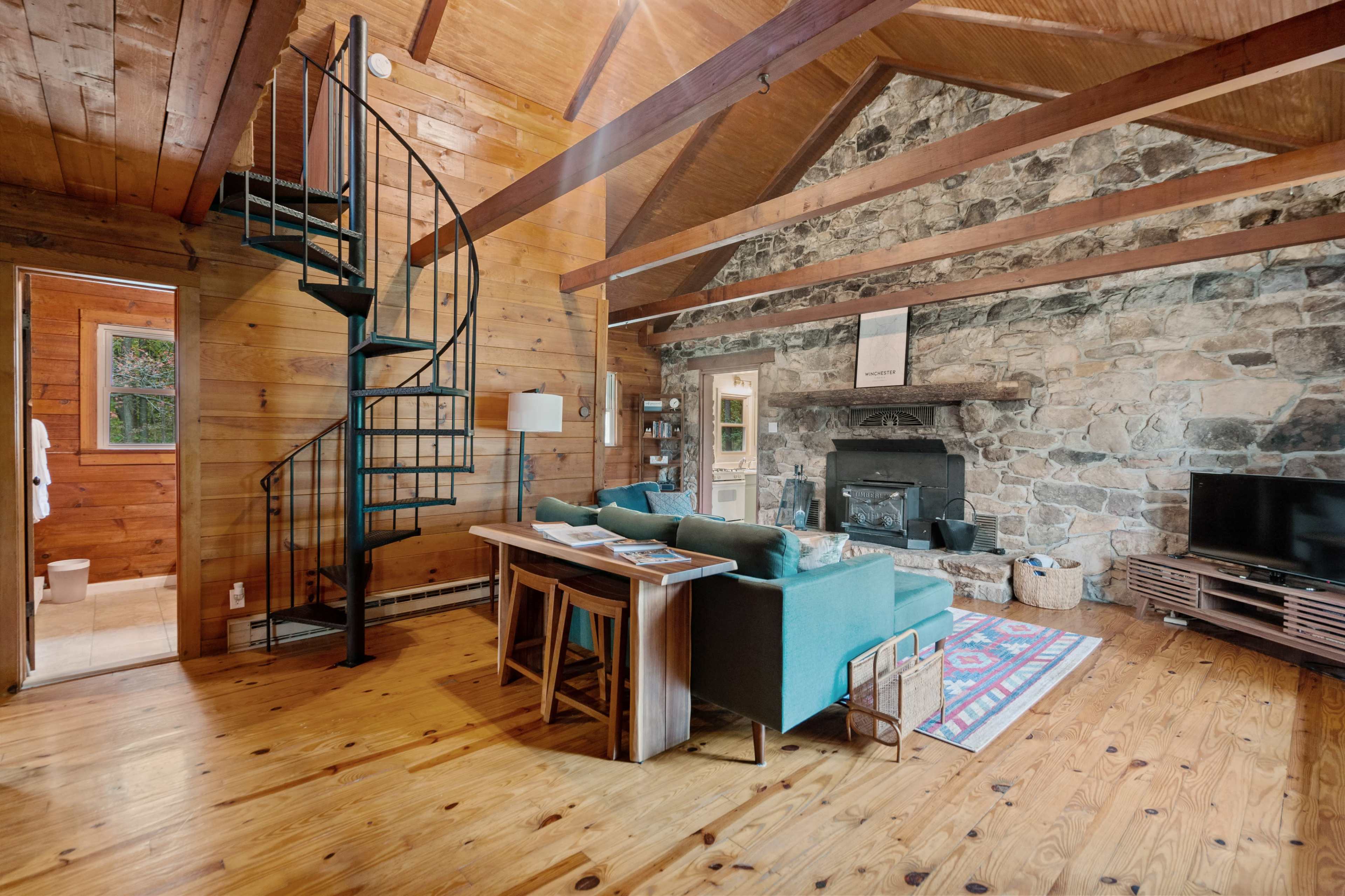 A cozy living area with a stone fireplace, wooden beams, a spiral staircase, and a turquoise sofa facing a television.