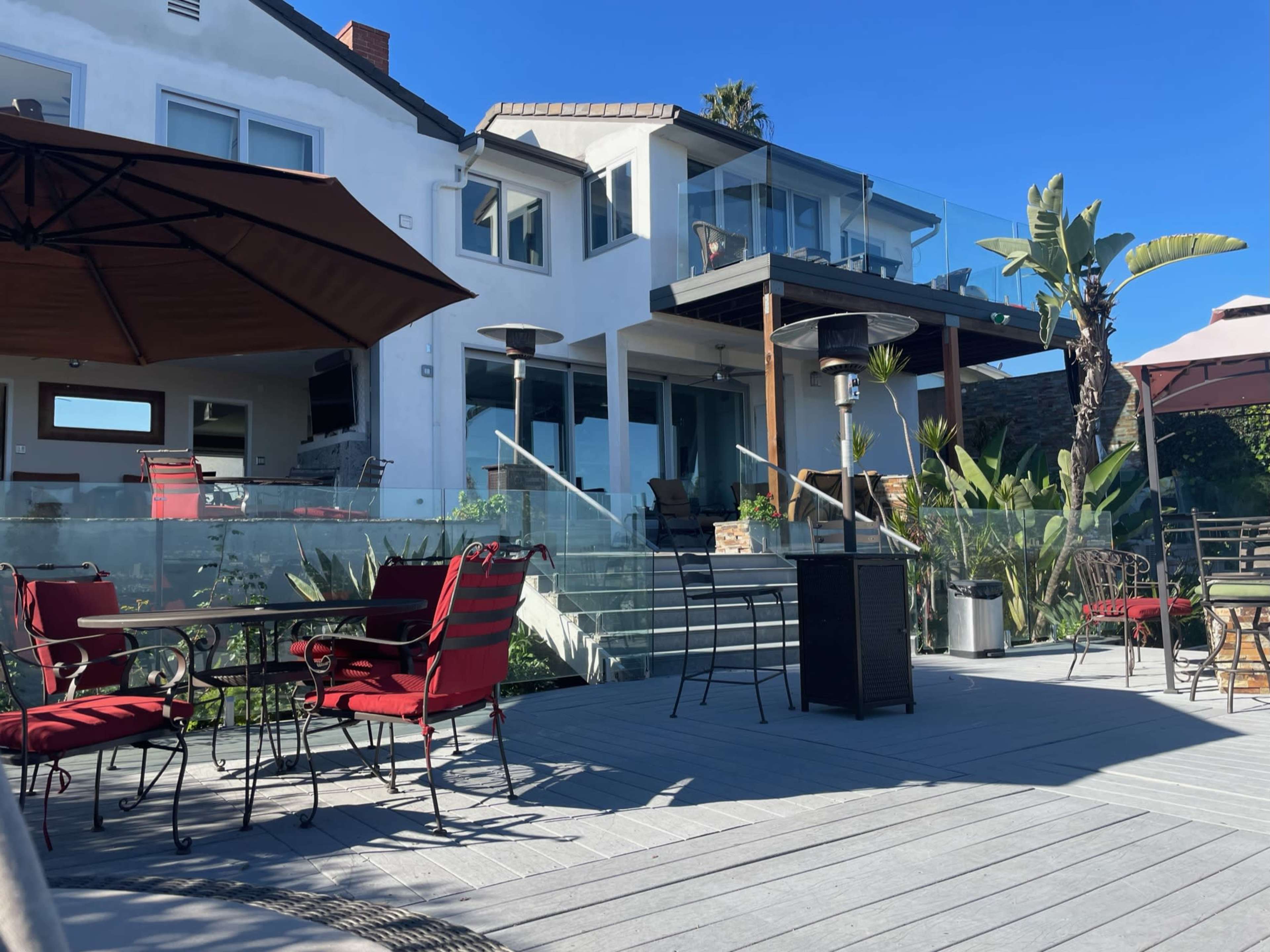 A modern outdoor patio with red chairs and tables is situated in front of a contemporary house featuring a glass balcony and palm trees.