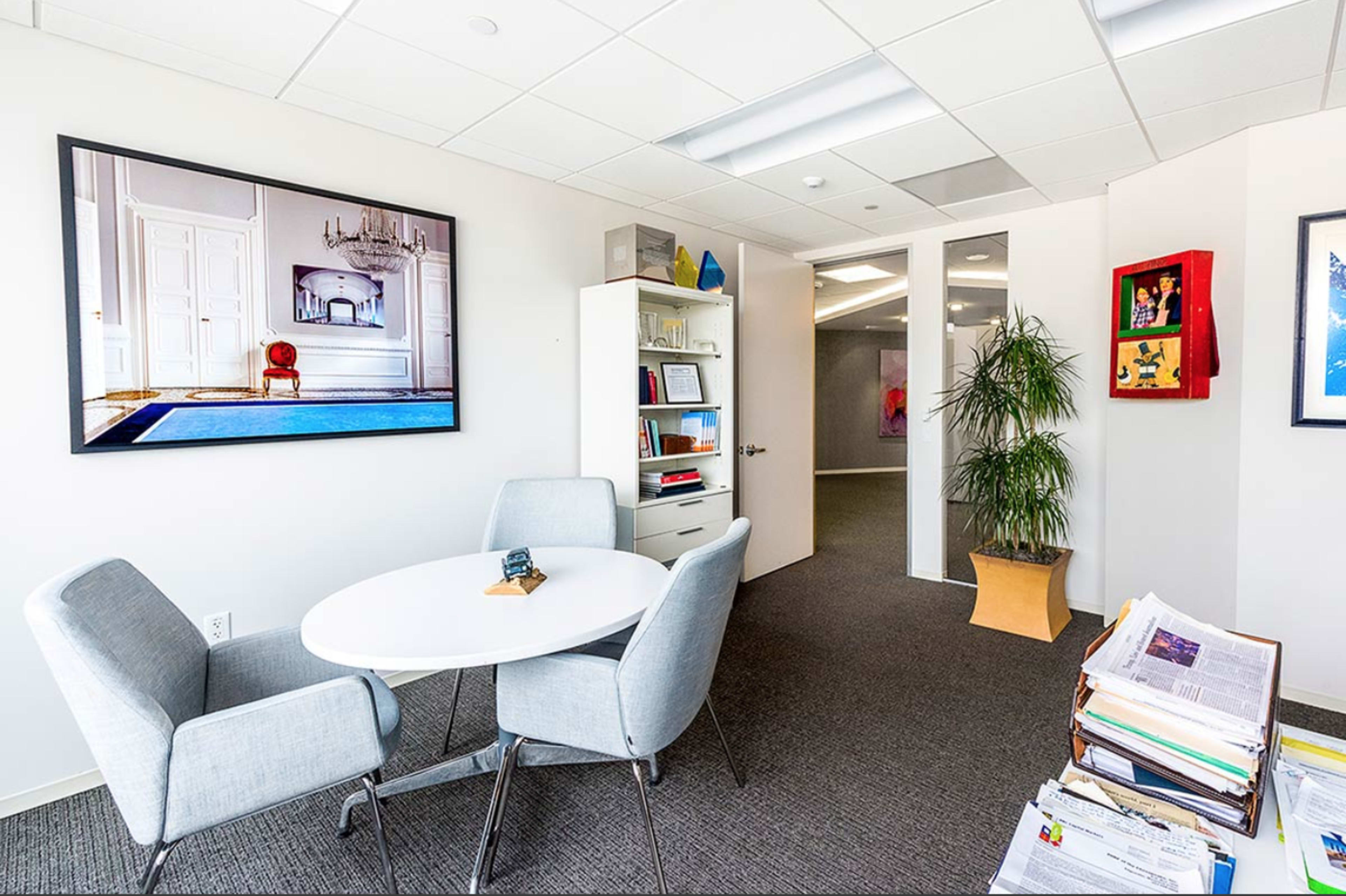 An office space featuring a round table with two chairs, a bookshelf, a large framed photo on the wall, and a potted plant.