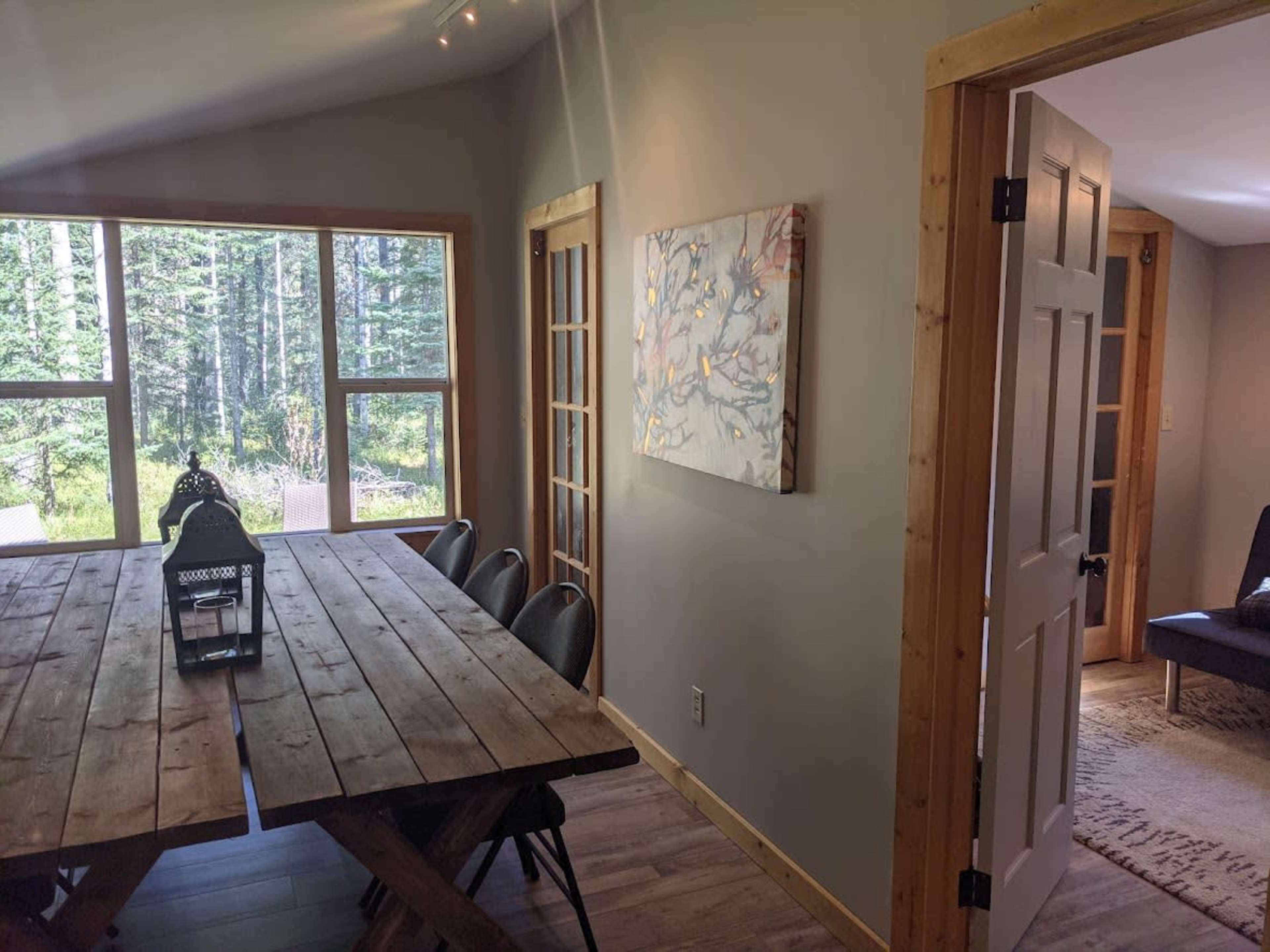 A dining area features a wooden table and chairs, with large windows displaying a view of a forested area outside.