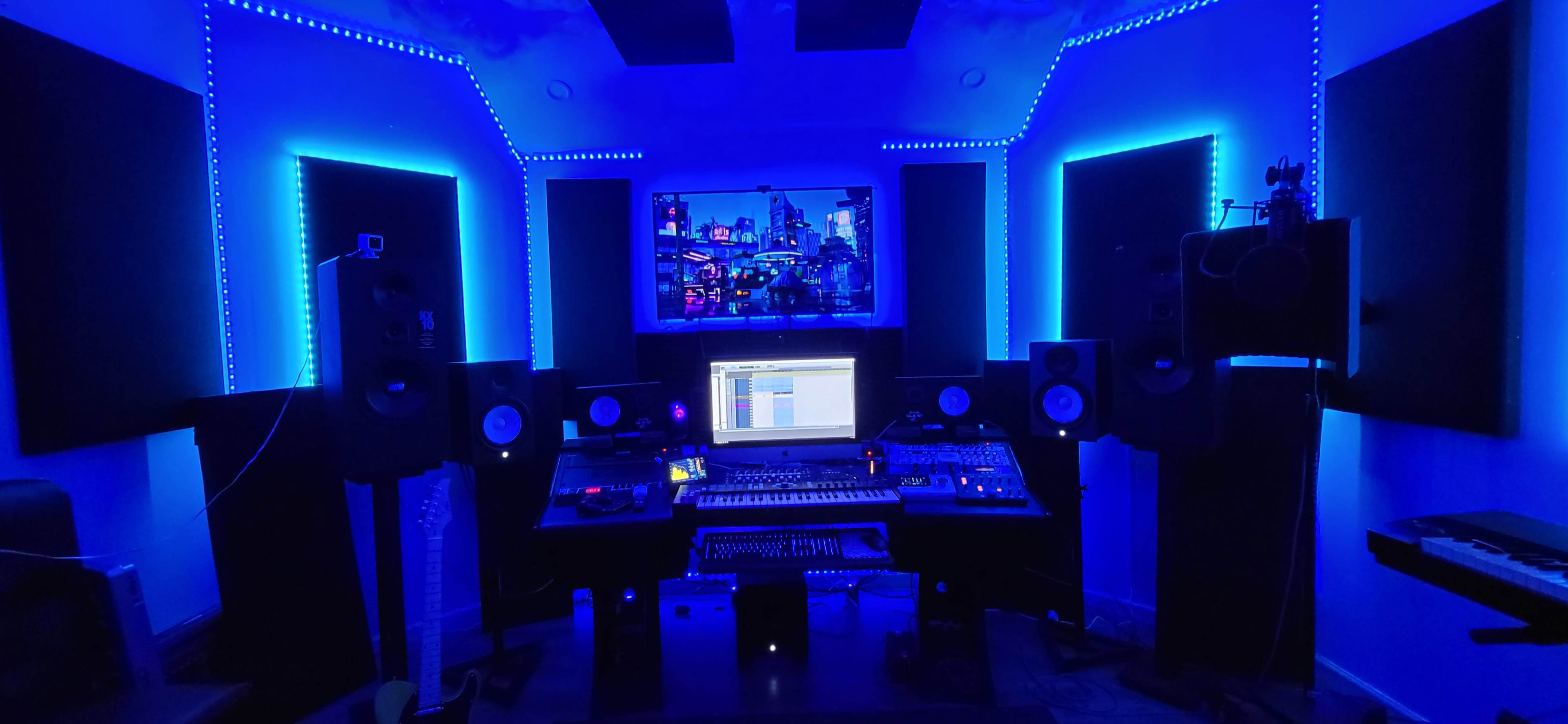 A music studio with blue LED lighting, featuring a computer desk with audio equipment, speakers, and a large screen displaying music software.