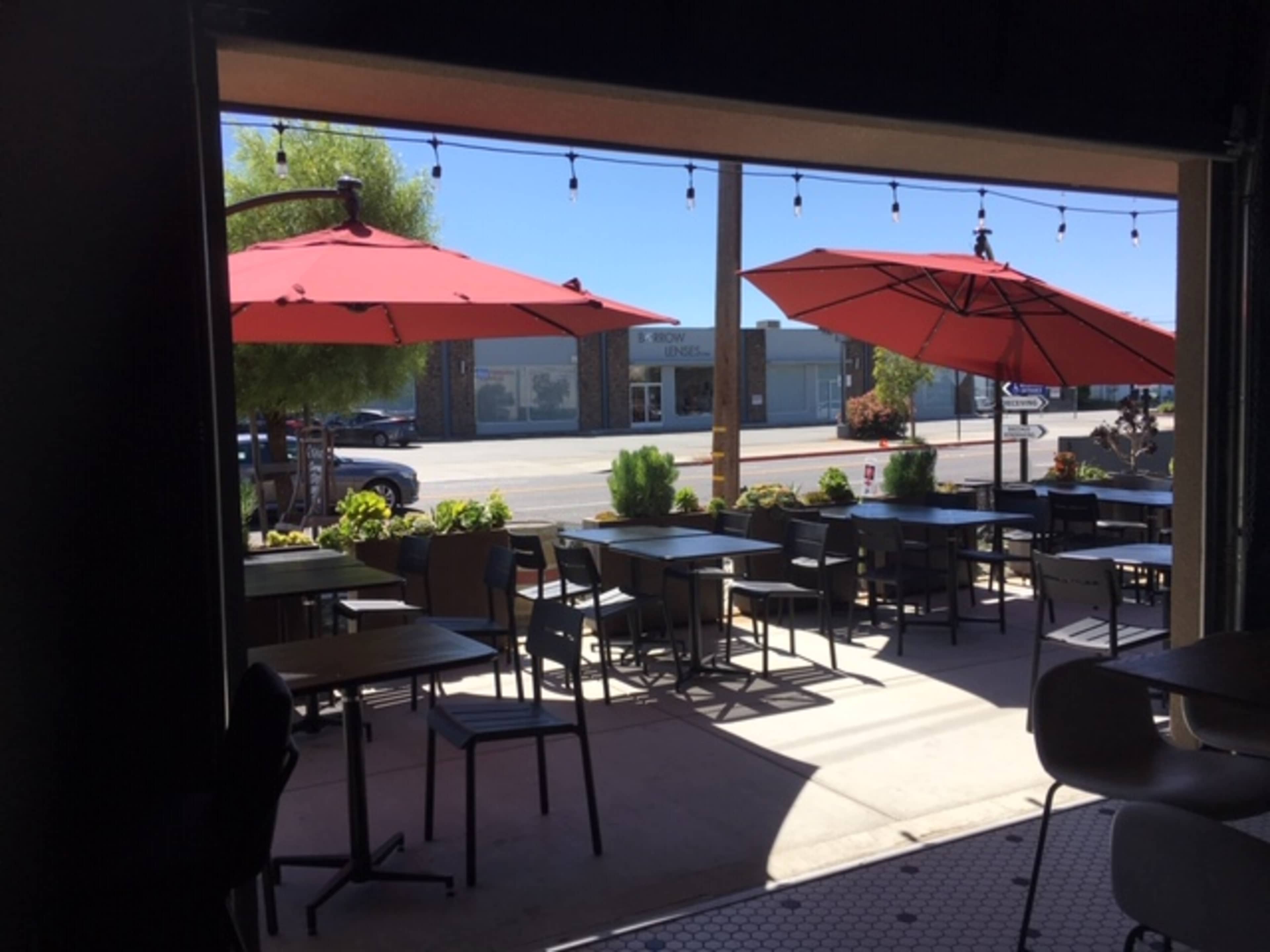 An outdoor dining area with tables and red umbrellas, facing a street lined with buildings.