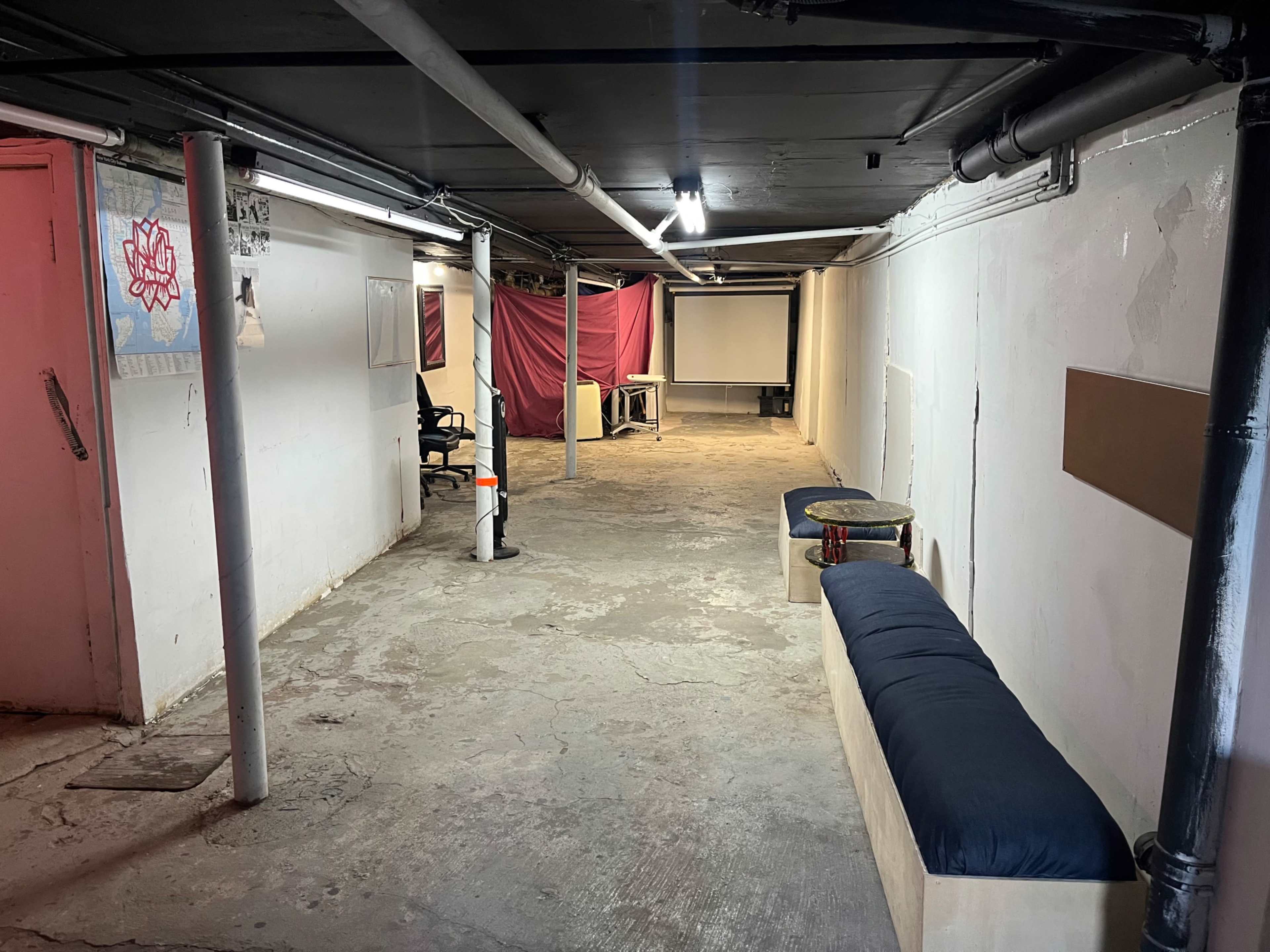 The image shows a dimly lit basement with concrete flooring, a pink curtain at the far end, a projector screen, a chair, and a long bench along one wall.