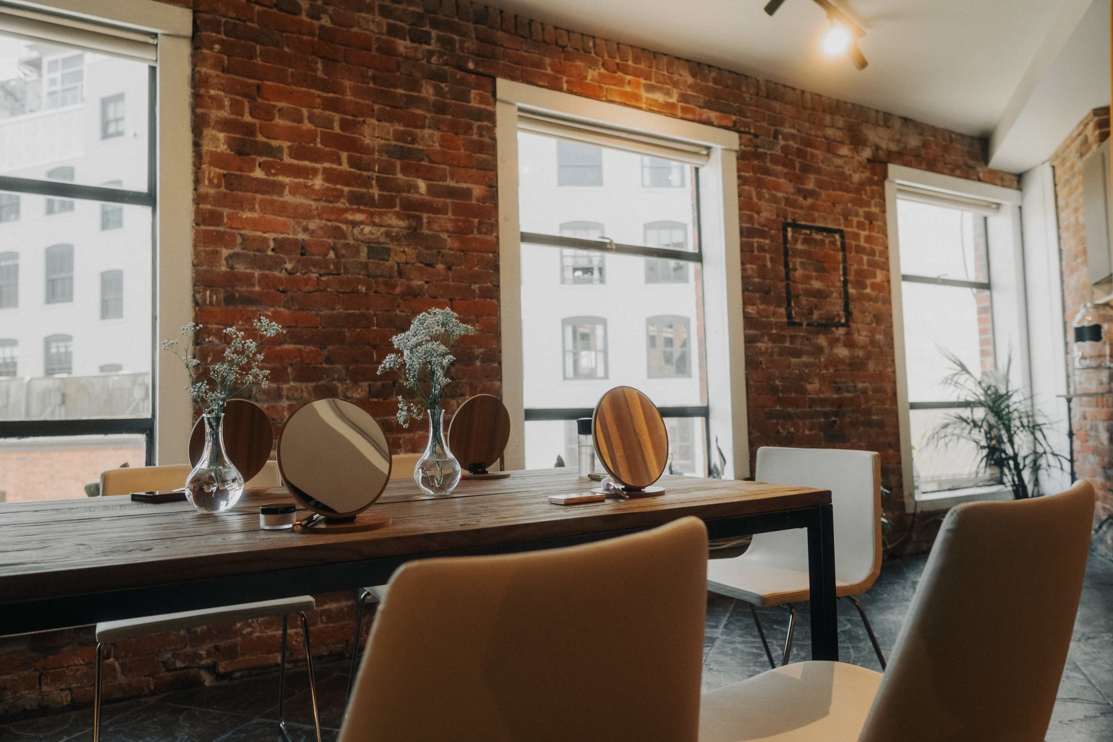 A modern interior with a wooden dining table, several round mirrors, and white chairs against a backdrop of exposed brick walls and large windows.