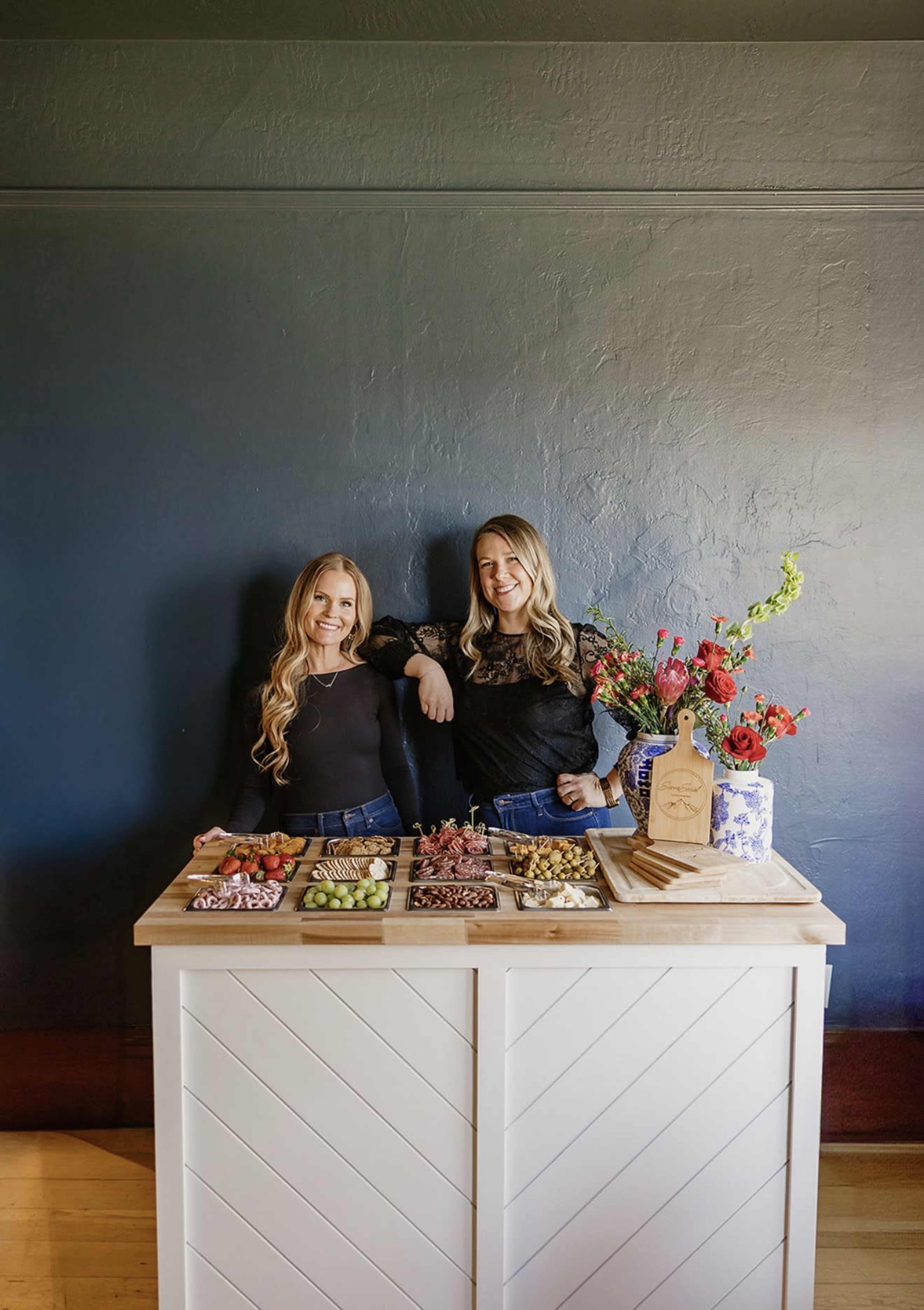 Two women stand beside a wooden table displaying an arrangement of food items, with a decorative floral centerpiece and a solid colored wall in the background.