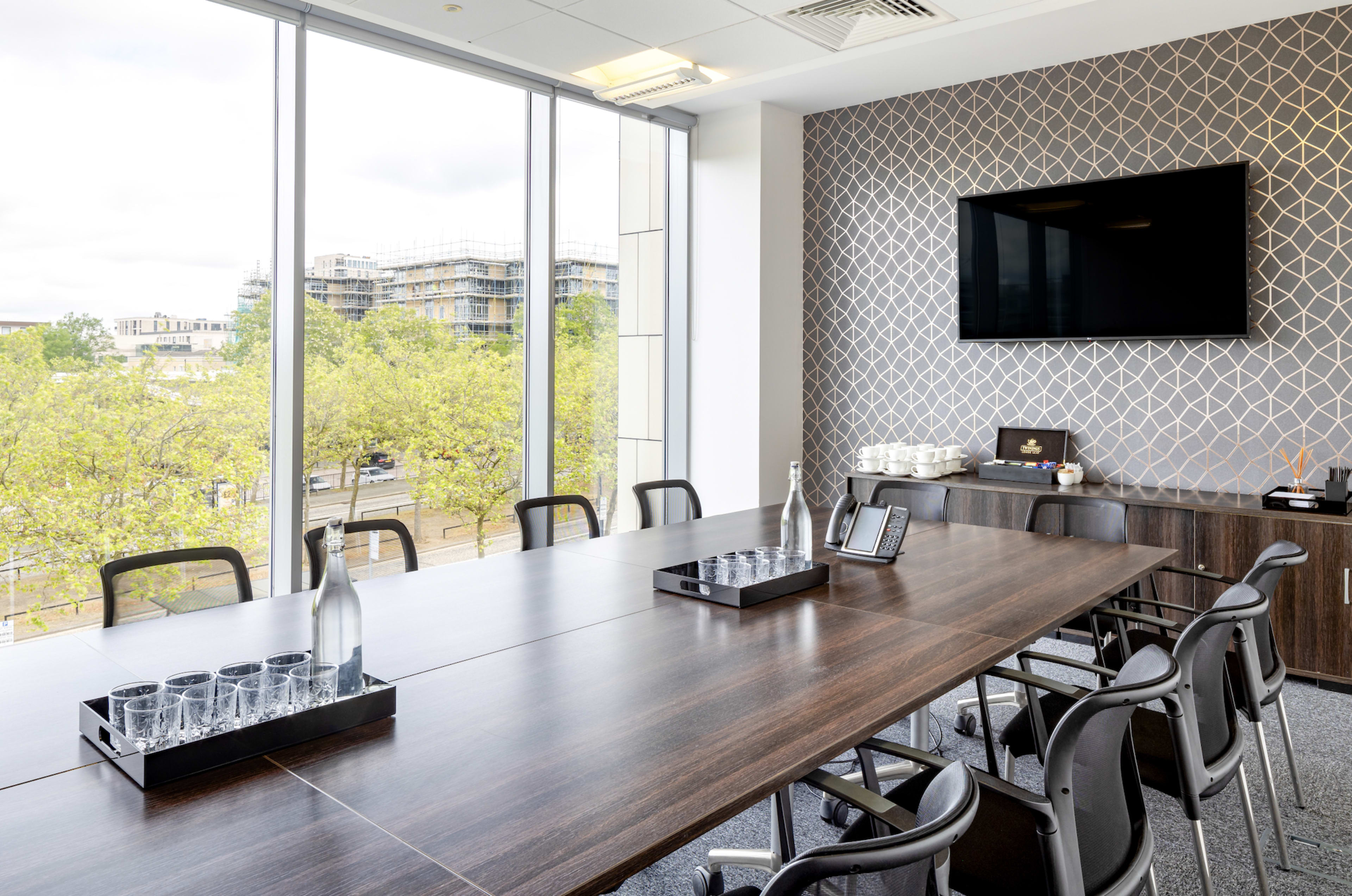 A modern conference room features a large wooden table surrounded by black chairs, with glassware and water bottles arranged on it, and large windows overlooking a tree-lined street.