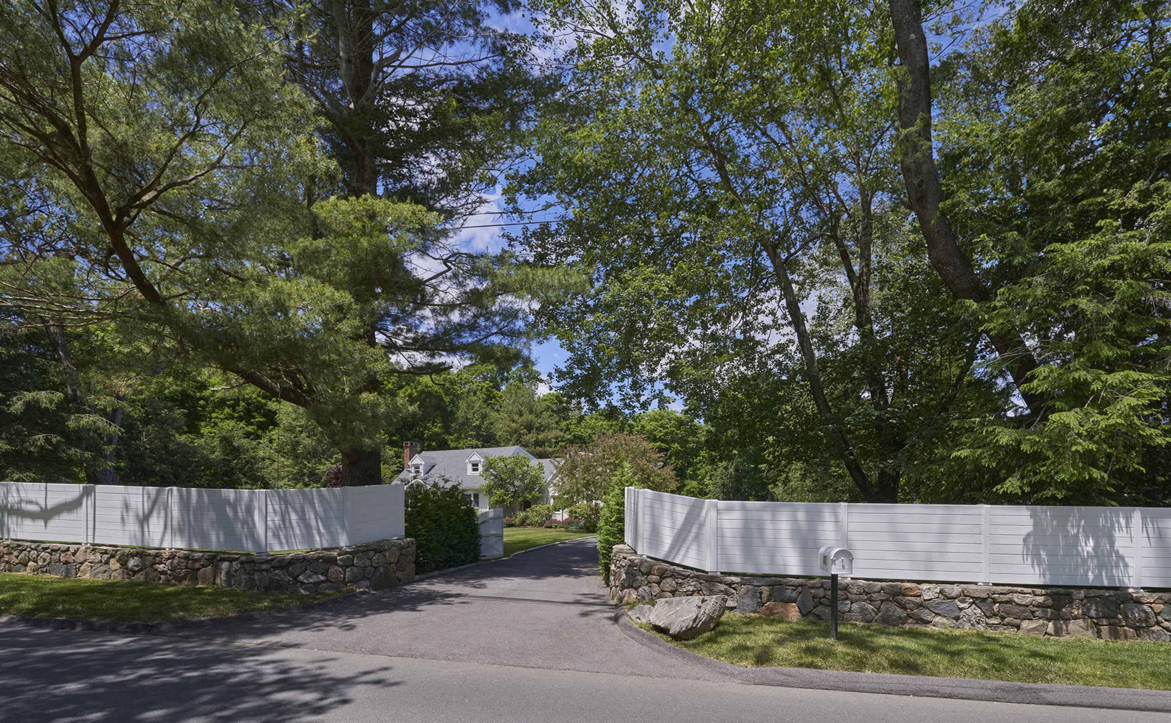 A winding driveway leads through a tree-lined entrance to a home behind white fences and stone walls.