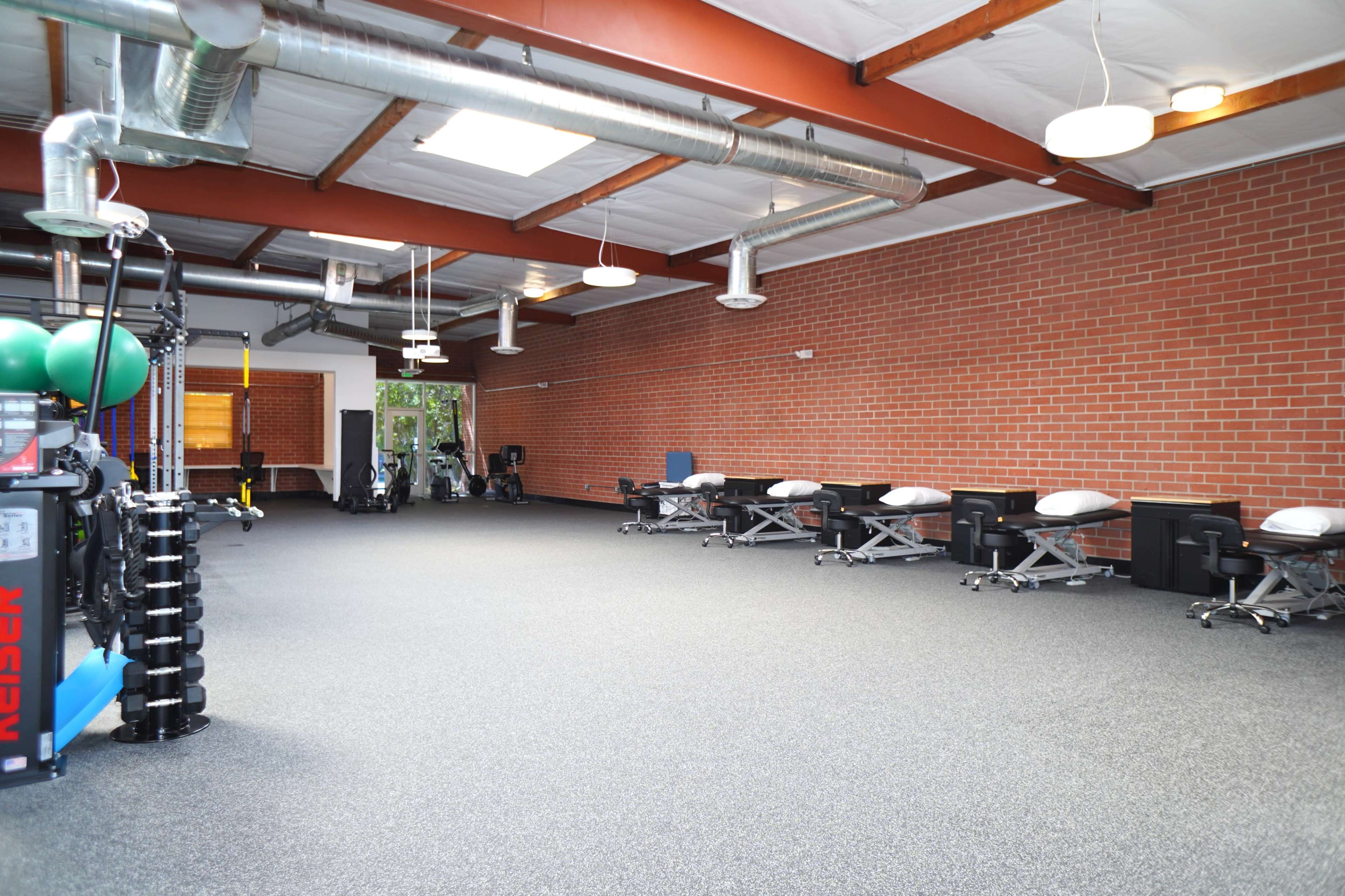 A spacious fitness studio with a rubber floor, brick walls, exercise equipment on the left, and treatment tables arranged on the right.