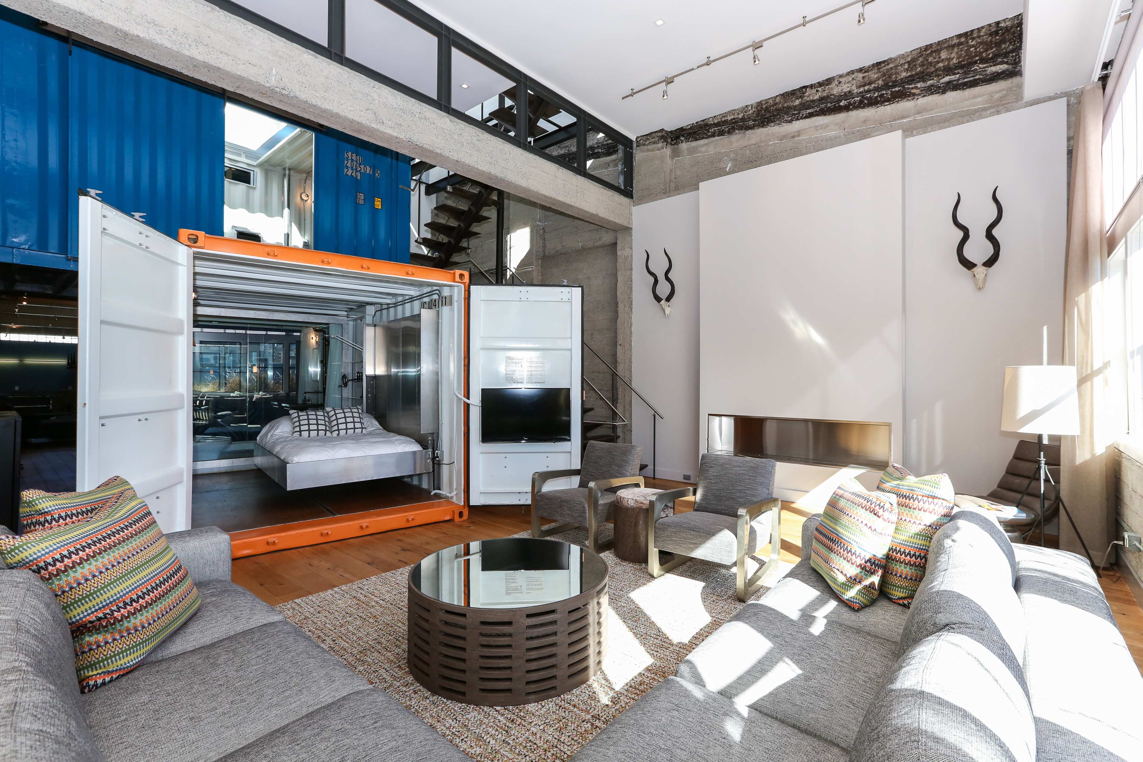 The image shows a modern living room featuring a shipping container transformed into a bedroom, with large windows, a sectional sofa, and decorative accents.