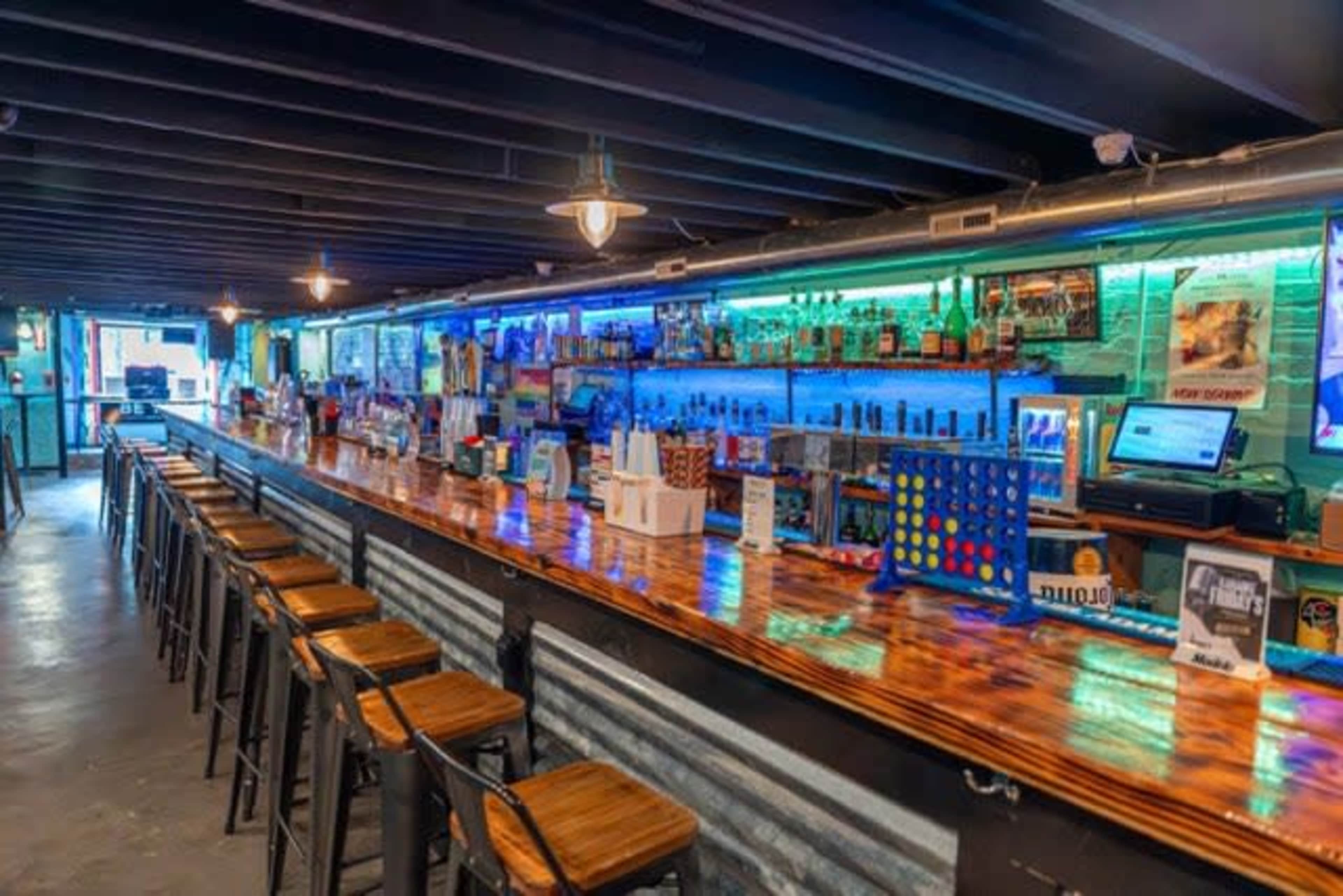 A long wooden bar is lined with metal stools, illuminated by colored lights and surrounded by shelves stocked with various bottles of liquor.