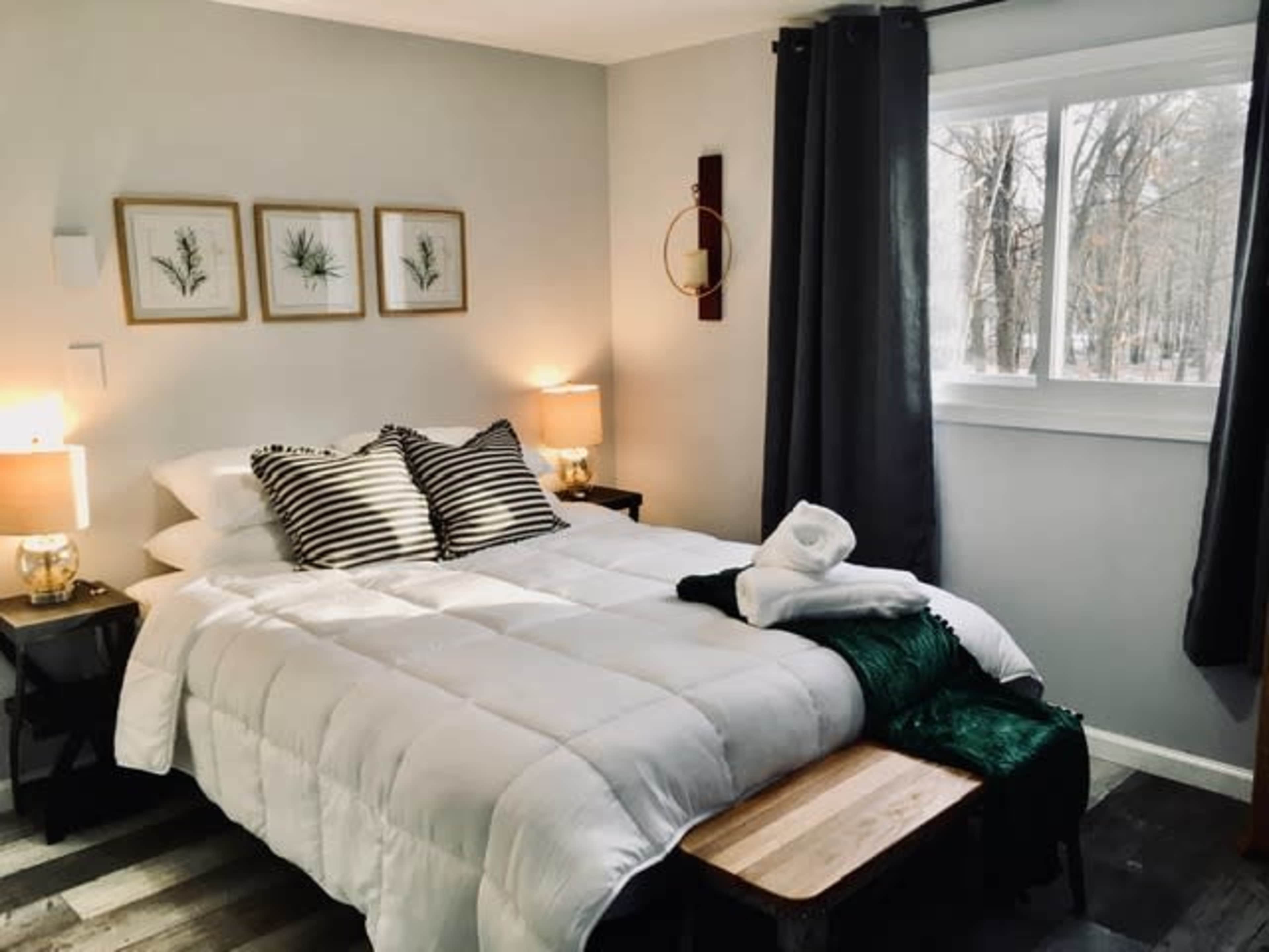 A neatly arranged bedroom featuring a bed with white bedding, framed botanical prints on the wall, and two bedside lamps next to the bed.