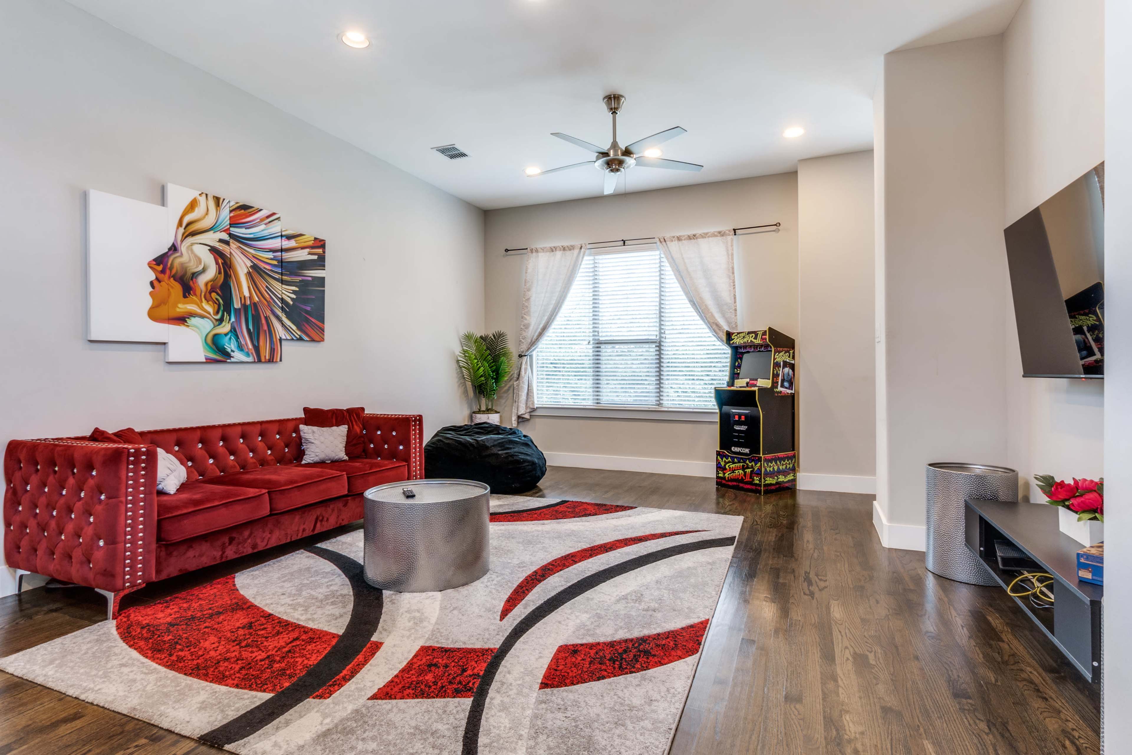 The room features a red tufted sofa, a round coffee table, a colorful area rug, and a corner with a tall arcade game machine.