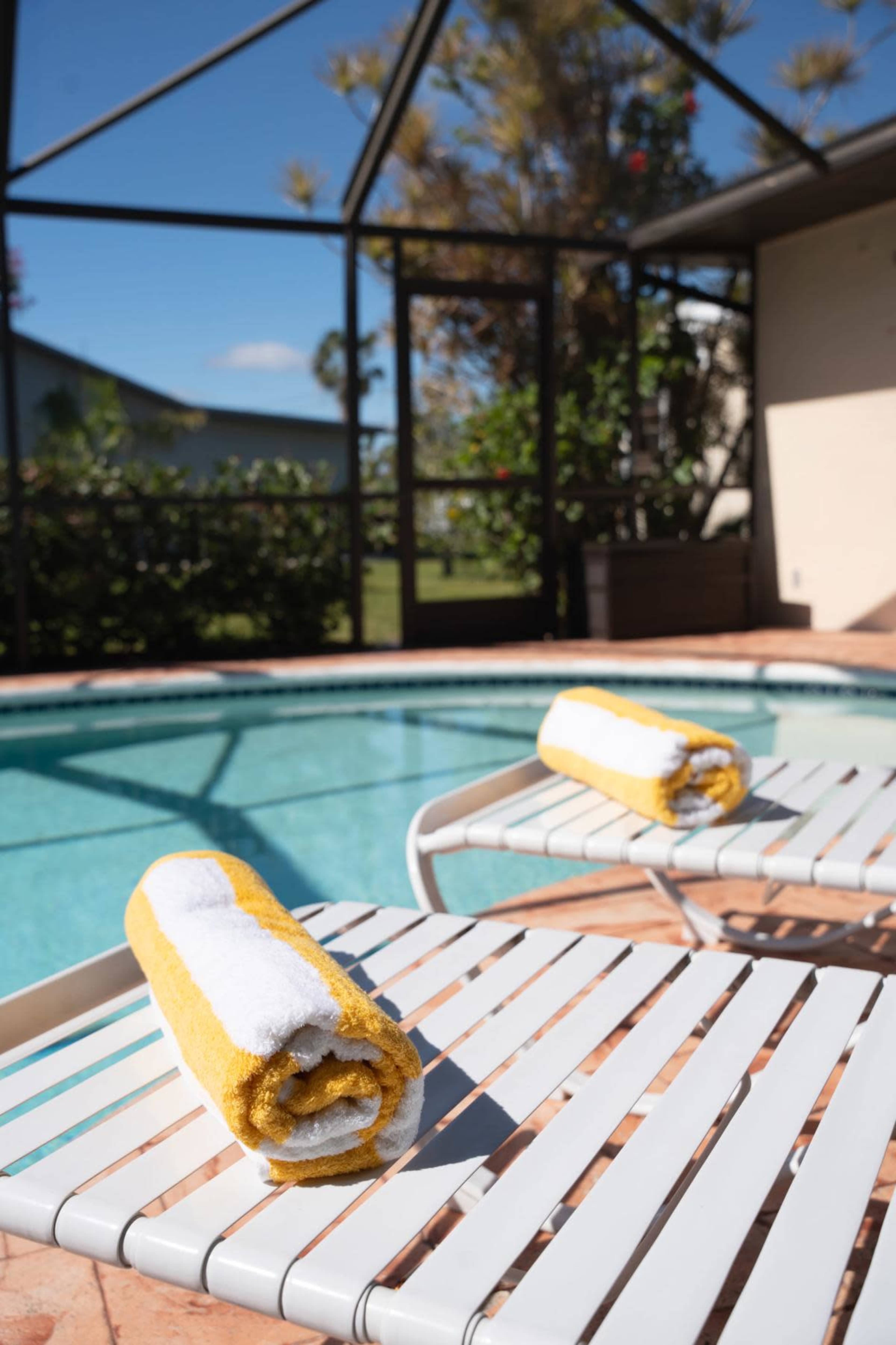 Two rolled towels sit on white lounge chairs beside a swimming pool enclosed by a screen.