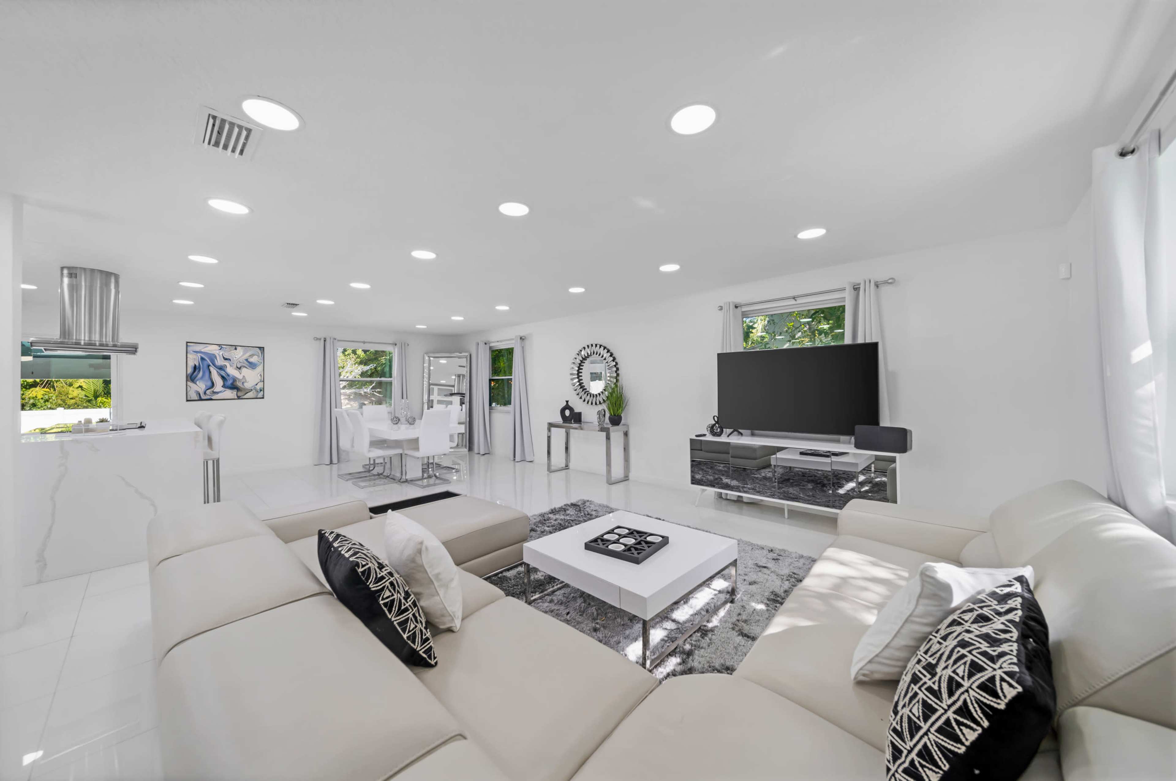 The image shows a modern living room with a large sectional sofa, a coffee table, and a wall-mounted television, illuminated by recessed lighting and natural light from windows.