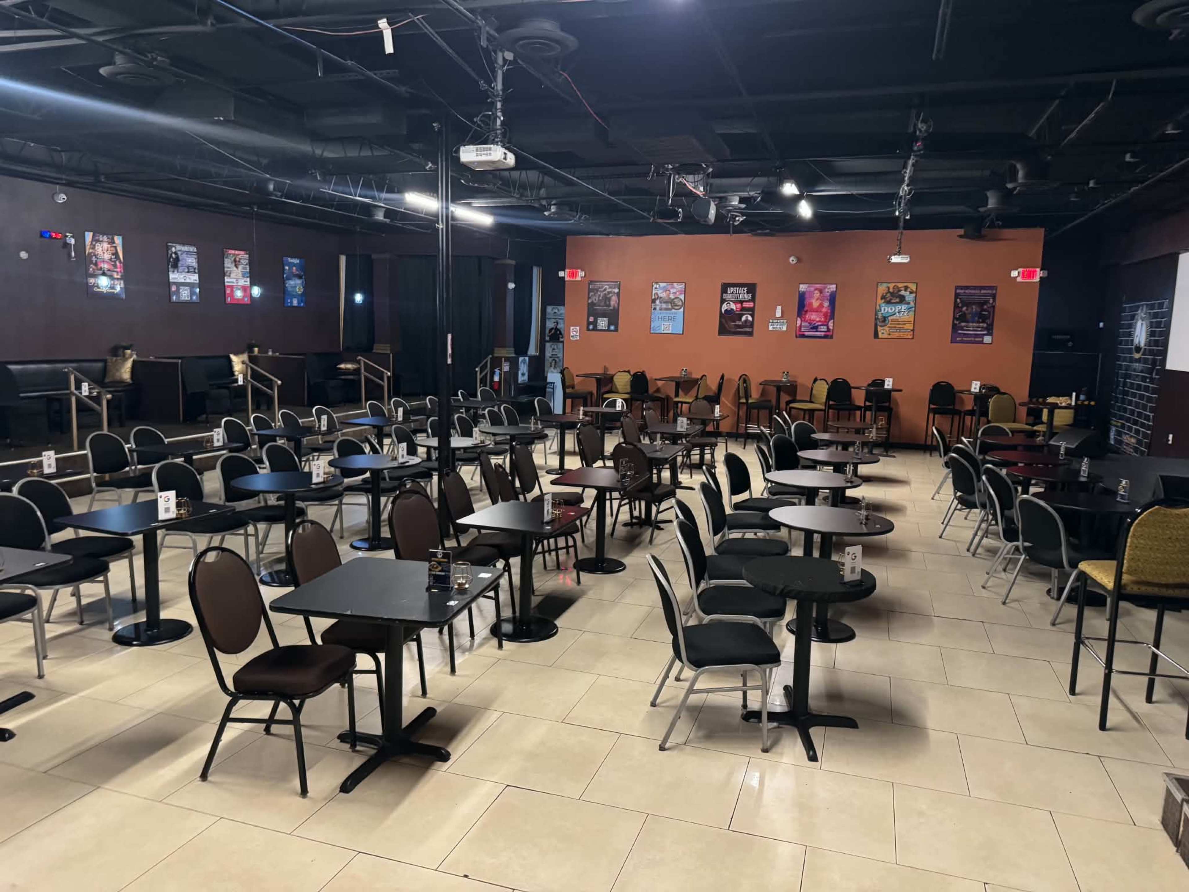 The image shows an empty, dimly lit event space with rows of tables and chairs arranged across the floor.
