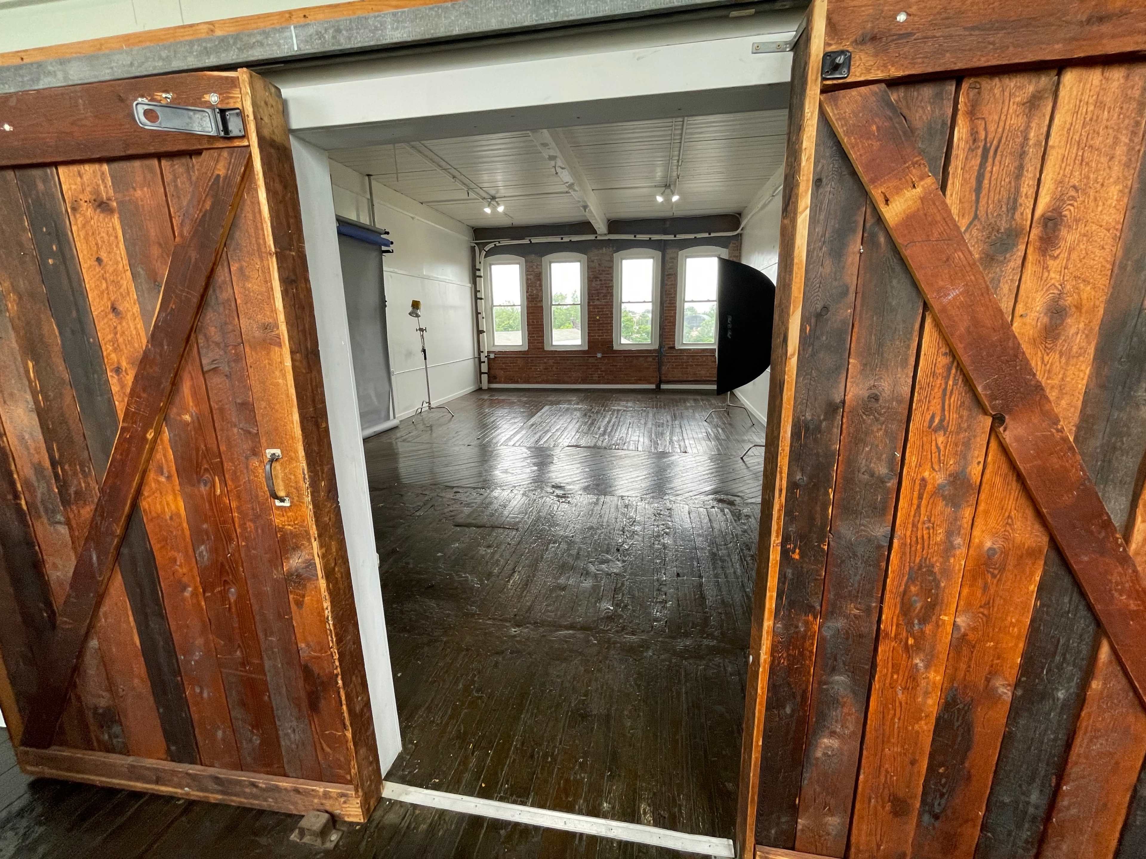 An open wooden double door leading into a bright room with large windows and wooden floors.