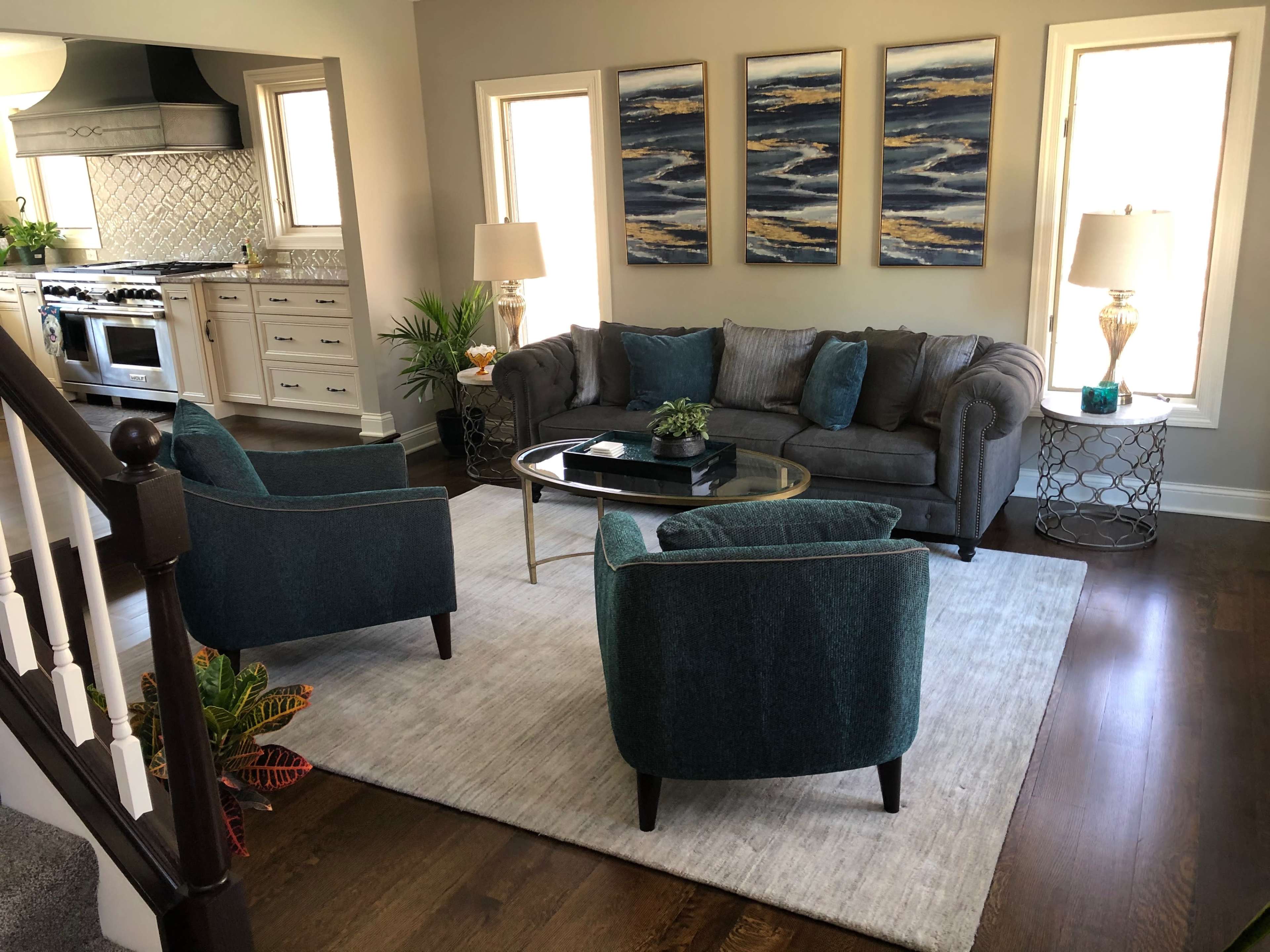 A modern living room features a gray sofa, two teal armchairs, a glass coffee table, and three abstract paintings on the wall.