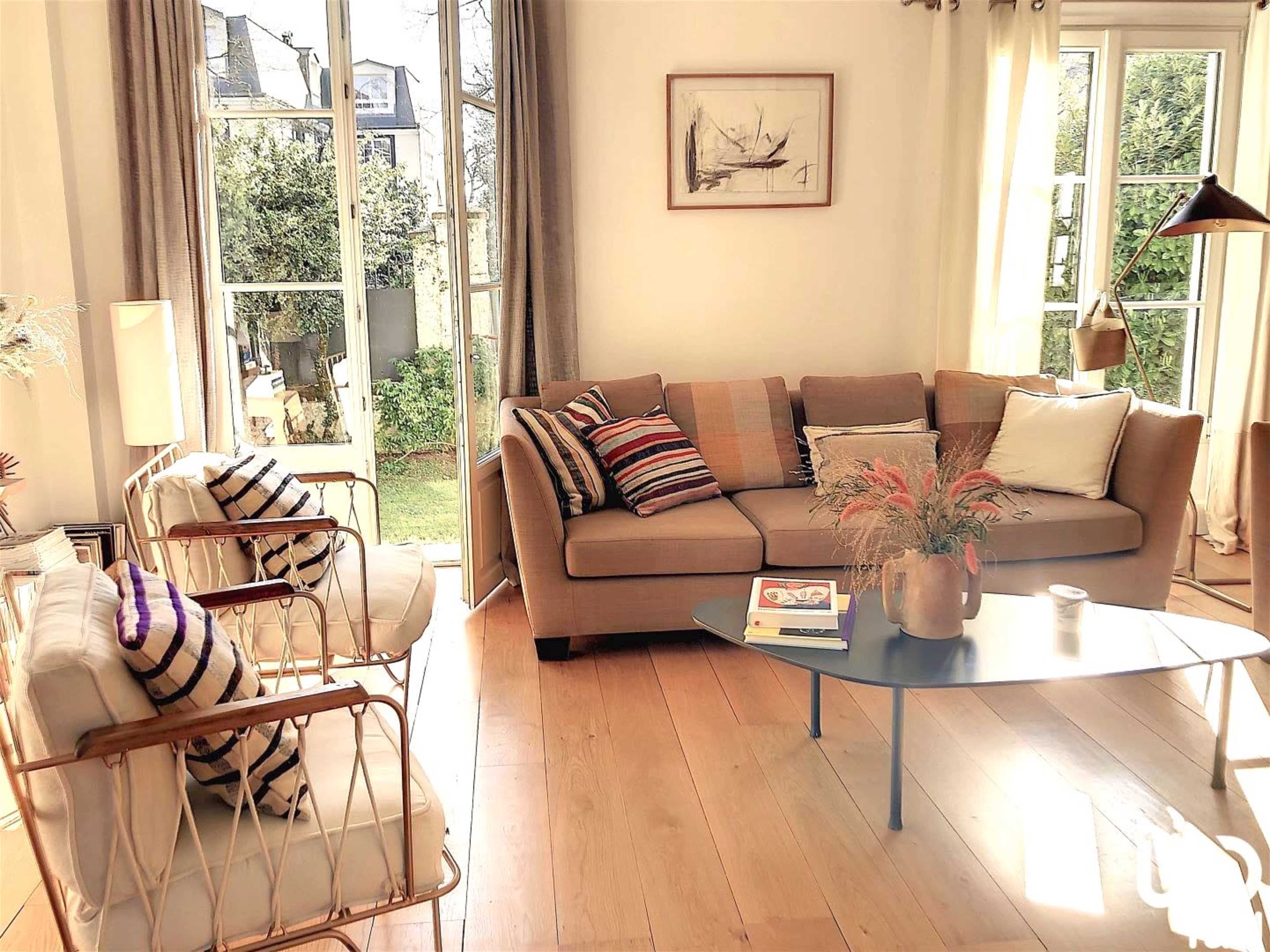 A cozy living room featuring a brown sofa with decorative cushions, two white chairs, a coffee table, and large windows that open to a garden.