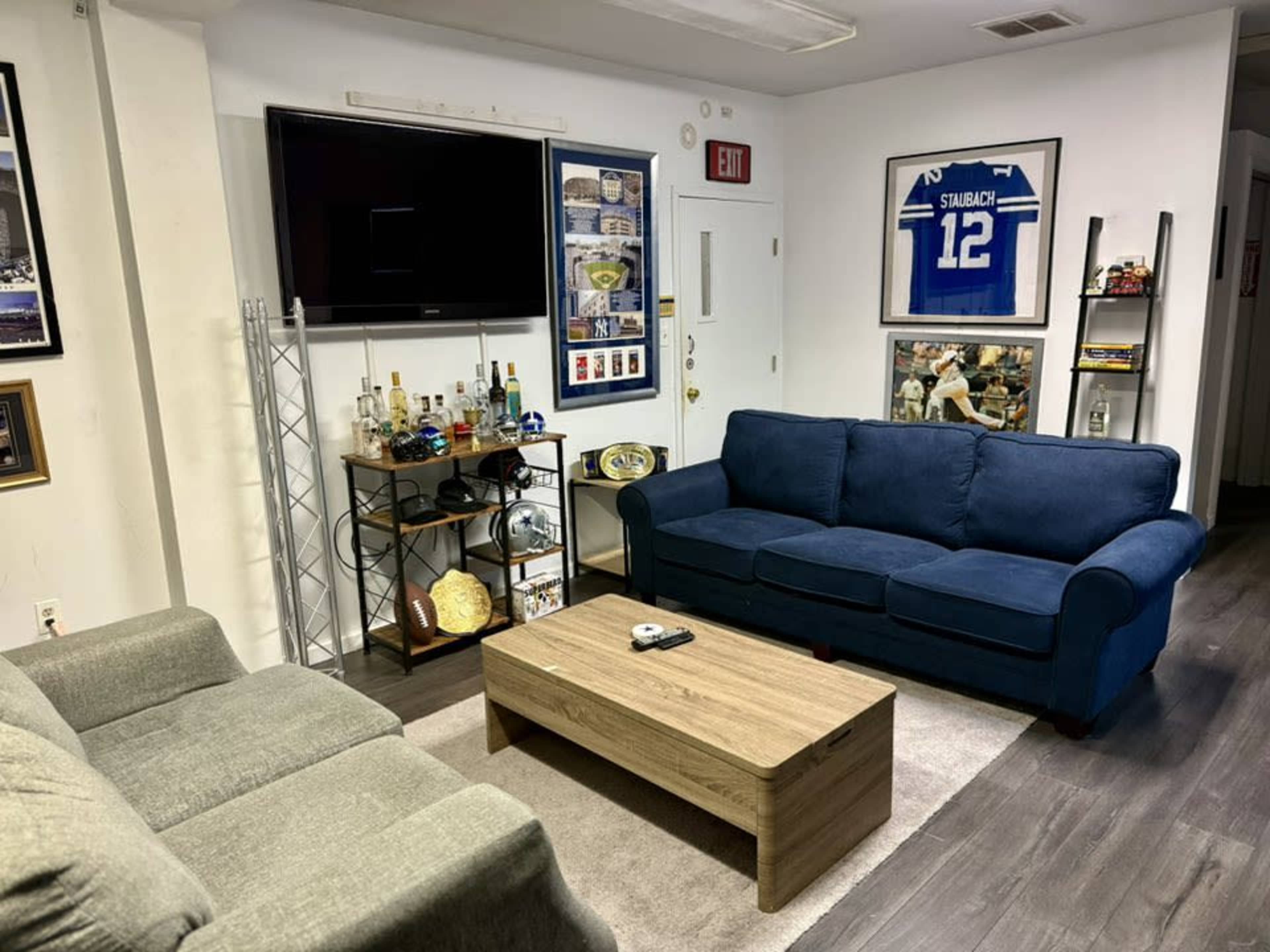 The image shows a living room featuring a blue couch, a gray couch, a wooden coffee table, a wall-mounted TV, and various decorations including shelves with awards and a sports jersey.