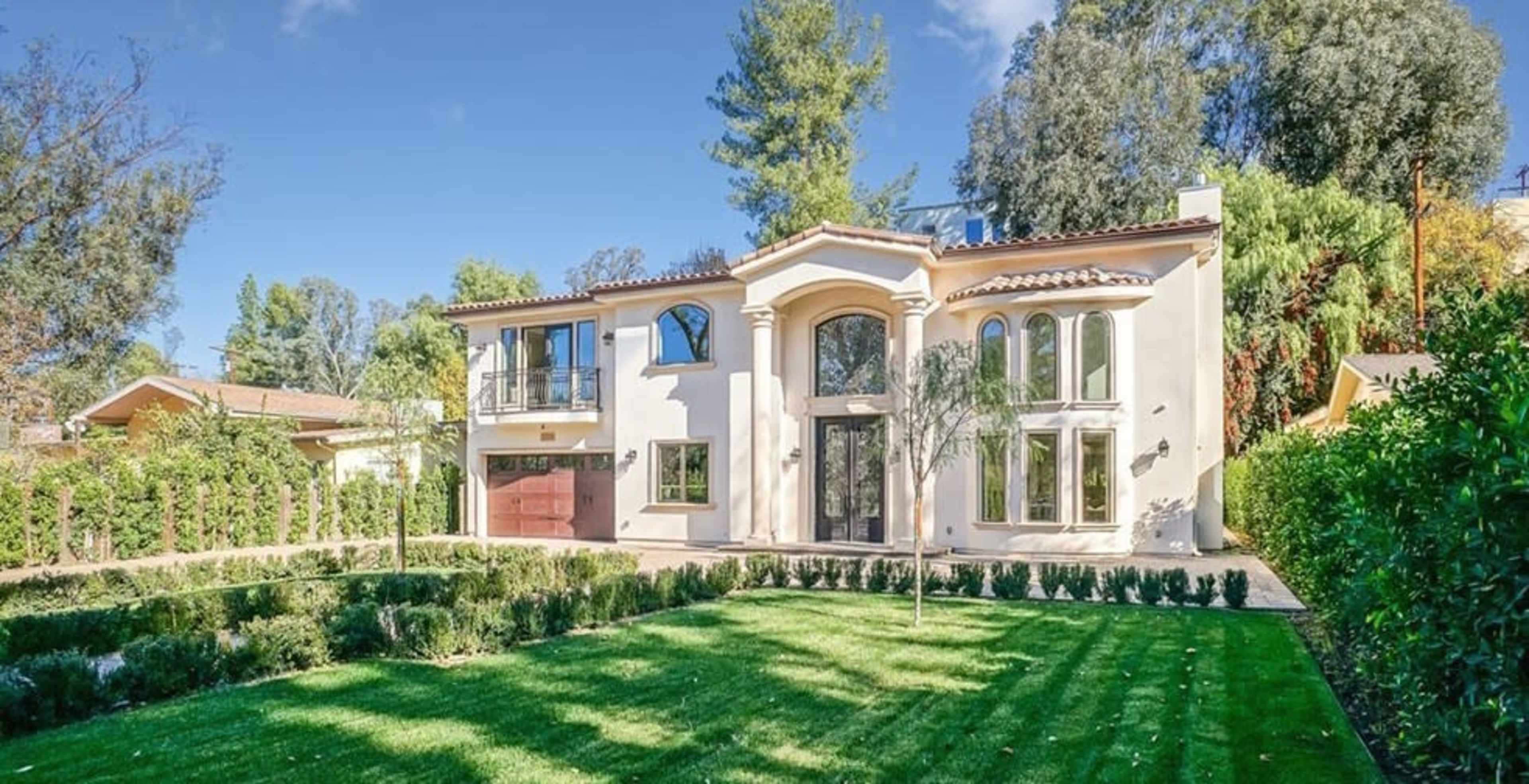 A large, two-story Mediterranean-style house with a red garage door and landscaped front yard is surrounded by trees and shrubs.