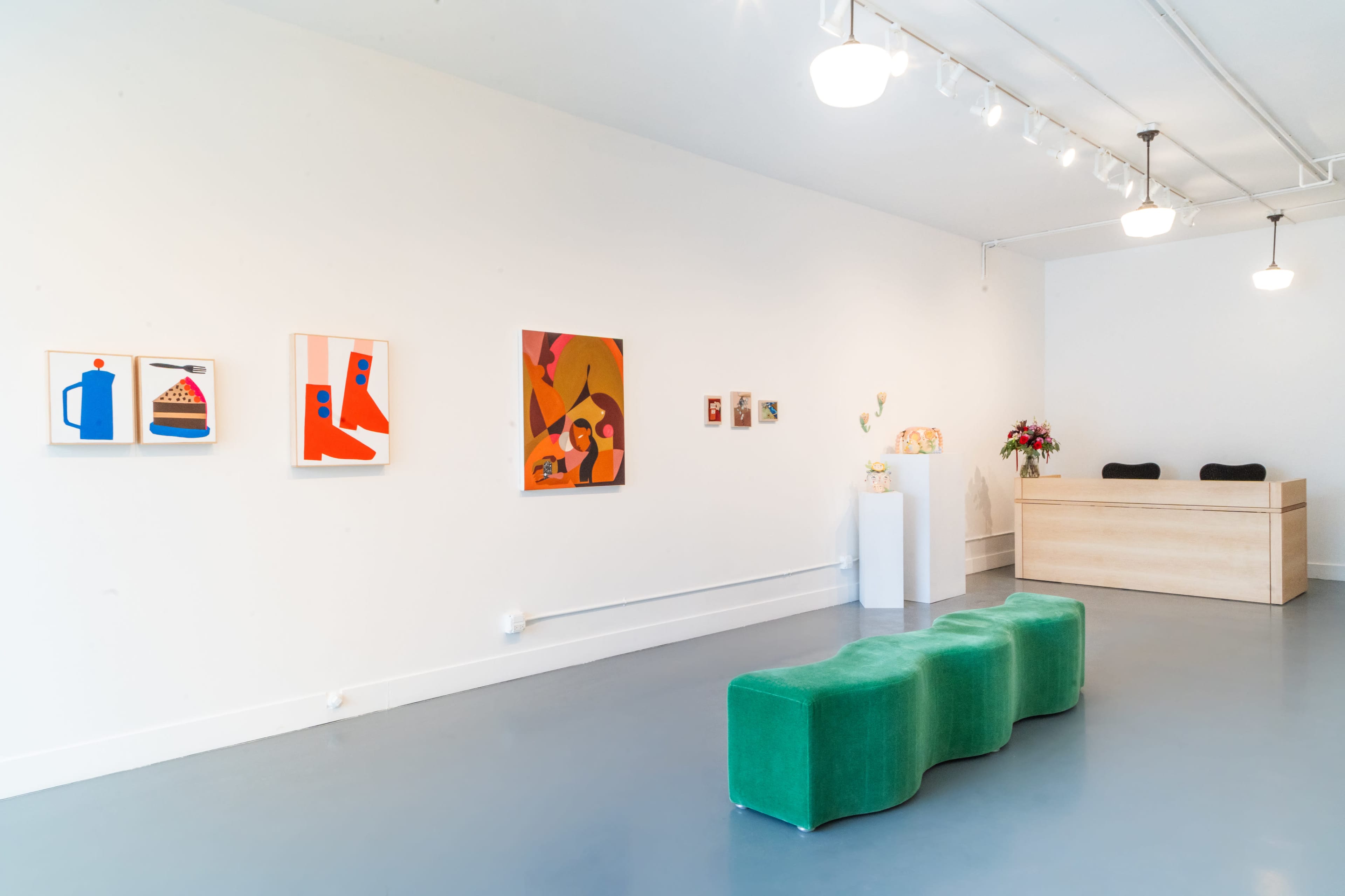 The image shows a modern art gallery featuring colorful paintings on white walls and a green seating area in front of a reception desk.