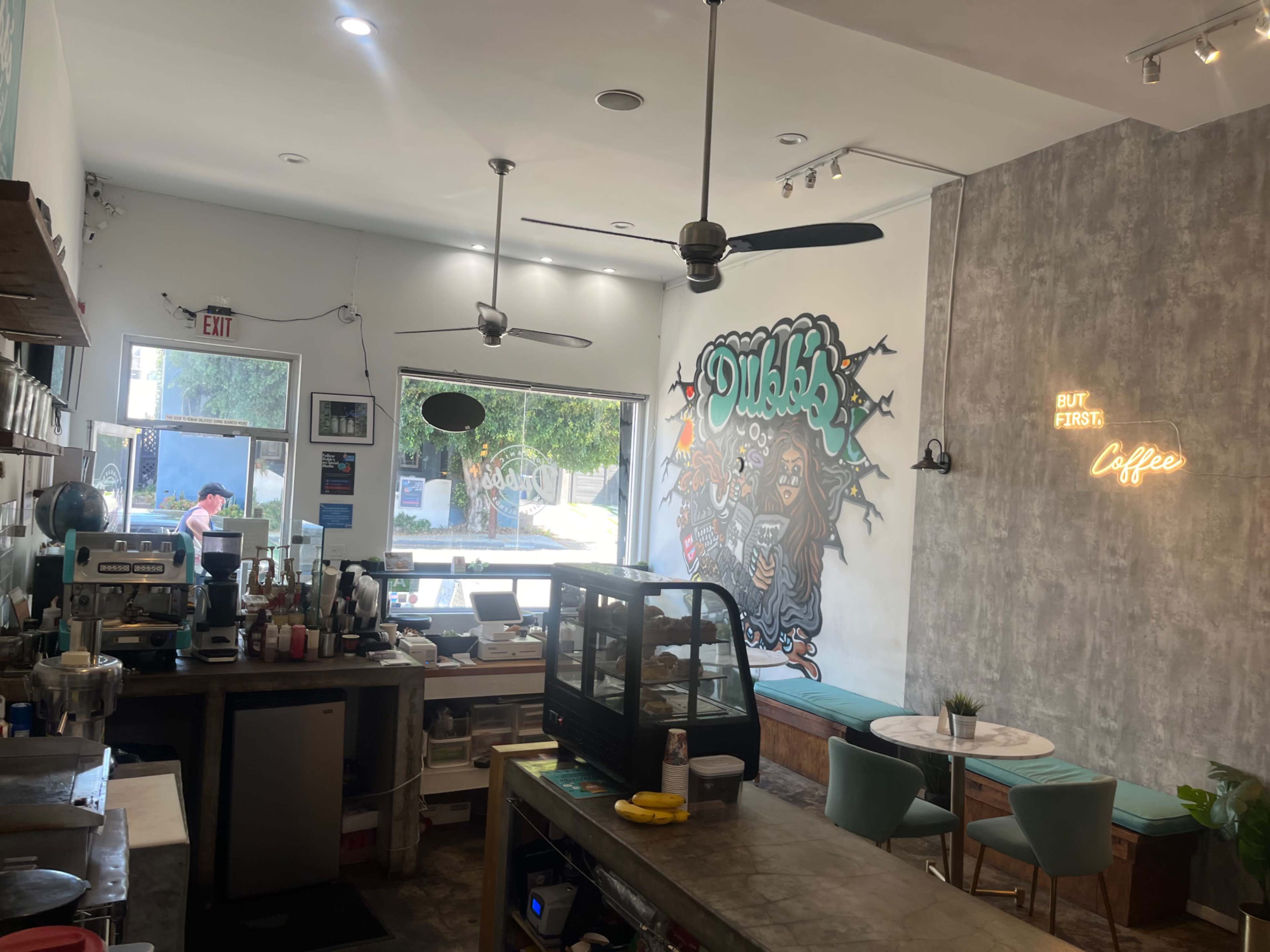 The image shows the interior of a café featuring a coffee counter, tables, and a mural on the wall with the words "But First, Coffee" illuminated in neon.
