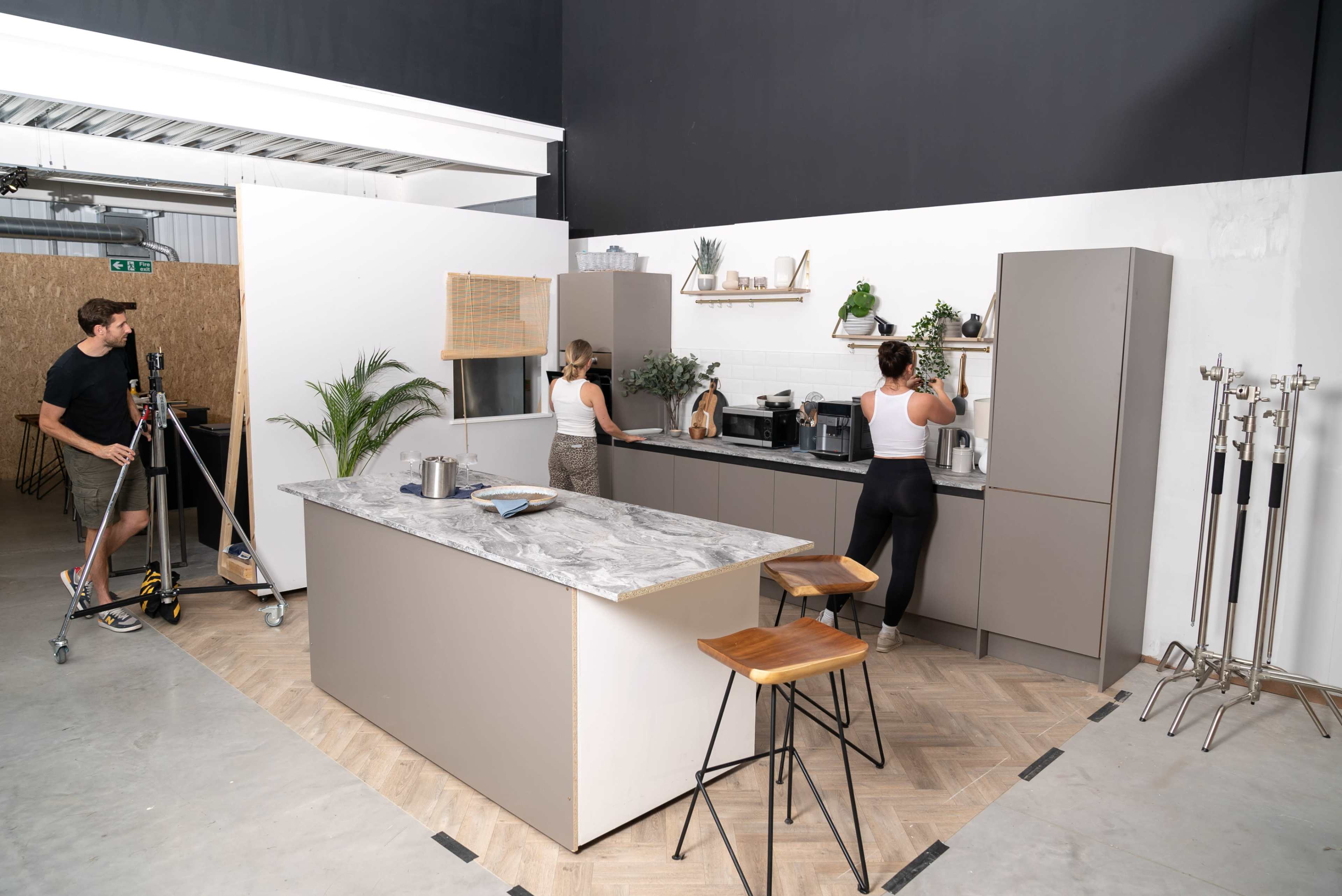 Two people are working in a modern kitchen with a central island while a third person adjusts a camera on a tripod.